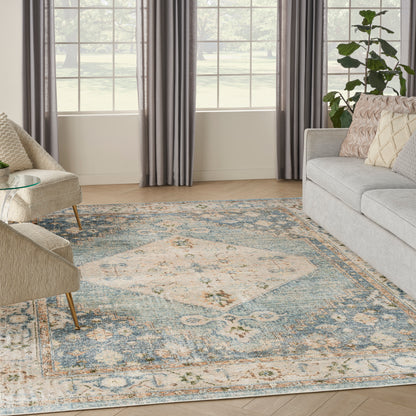 Cozy living room with grey curtains, beige upholstered armchairs, light sofa, blue floral rug, glass side table, potted plant.