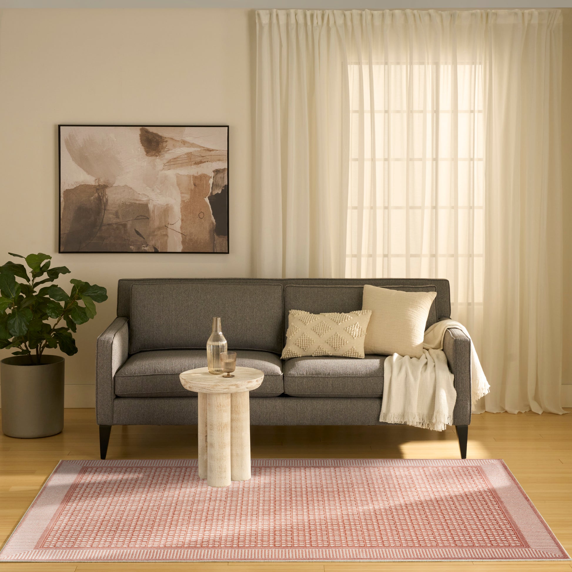 Gray fabric sofa with cream cushions and throw, round wooden side table, pink patterned rug, plant, sheer curtains.