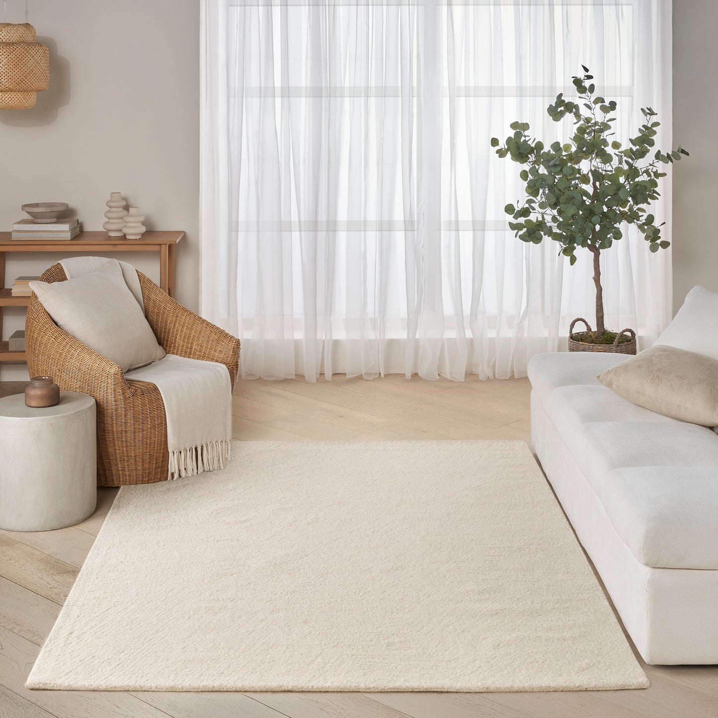 Neutral living room with cream rug, white sofa, wicker armchair, sheer curtains, potted tree, and light wood floors.