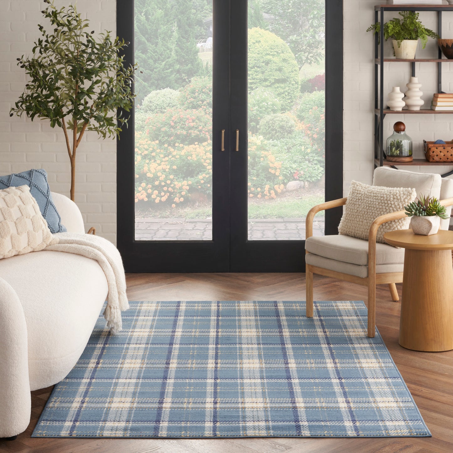 Blue plaid woven area rug in a sunlit living room with cream sofa, wooden chair, and glass doors to a lush garden.
