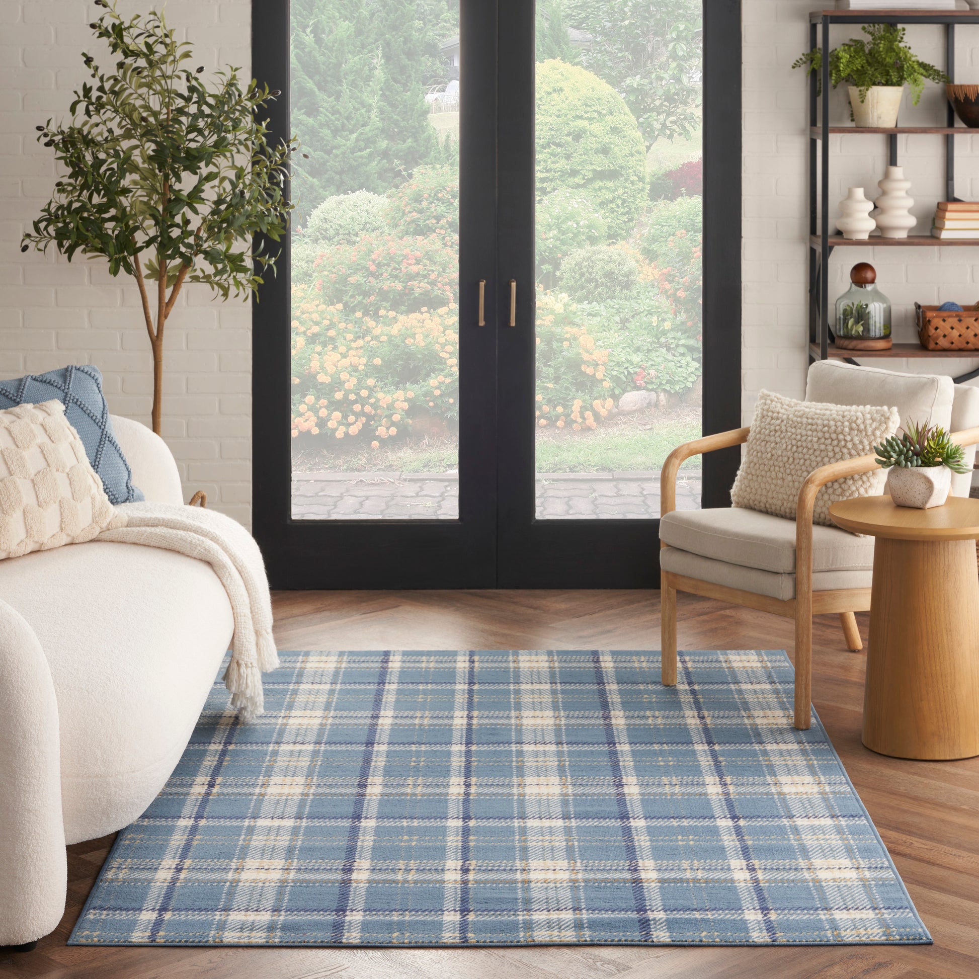 Blue plaid woven area rug in a sunlit living room with cream sofa, wooden chair, and glass doors to a lush garden.