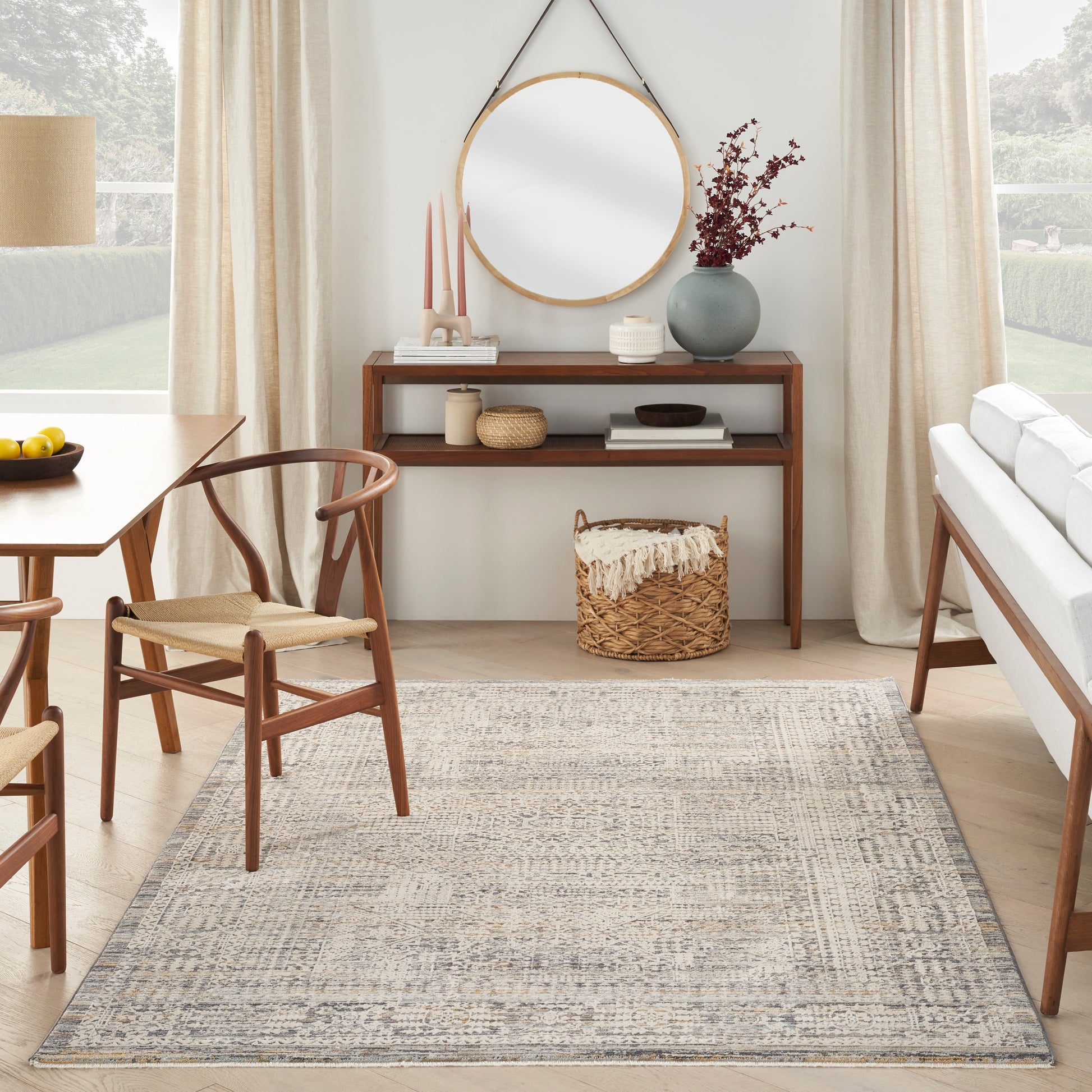Mid-century living room with wooden dining chairs and console, round wall mirror, vase of dried blooms, woven basket, and neutral rug.