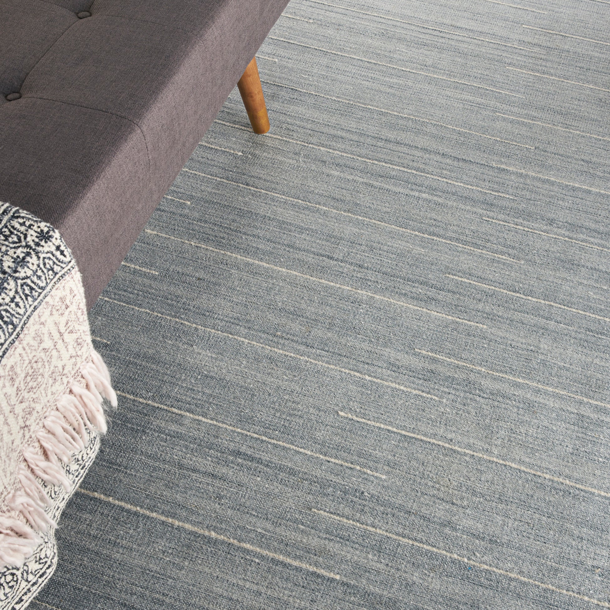 Gray fabric sofa corner with a wooden leg, blue-gray striped rug, and a fringed throw blanket.