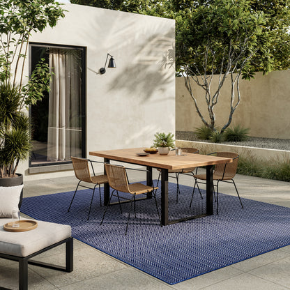 Outdoor dining set: wooden table with black metal legs, six wicker chairs, on a blue outdoor rug in a modern courtyard.