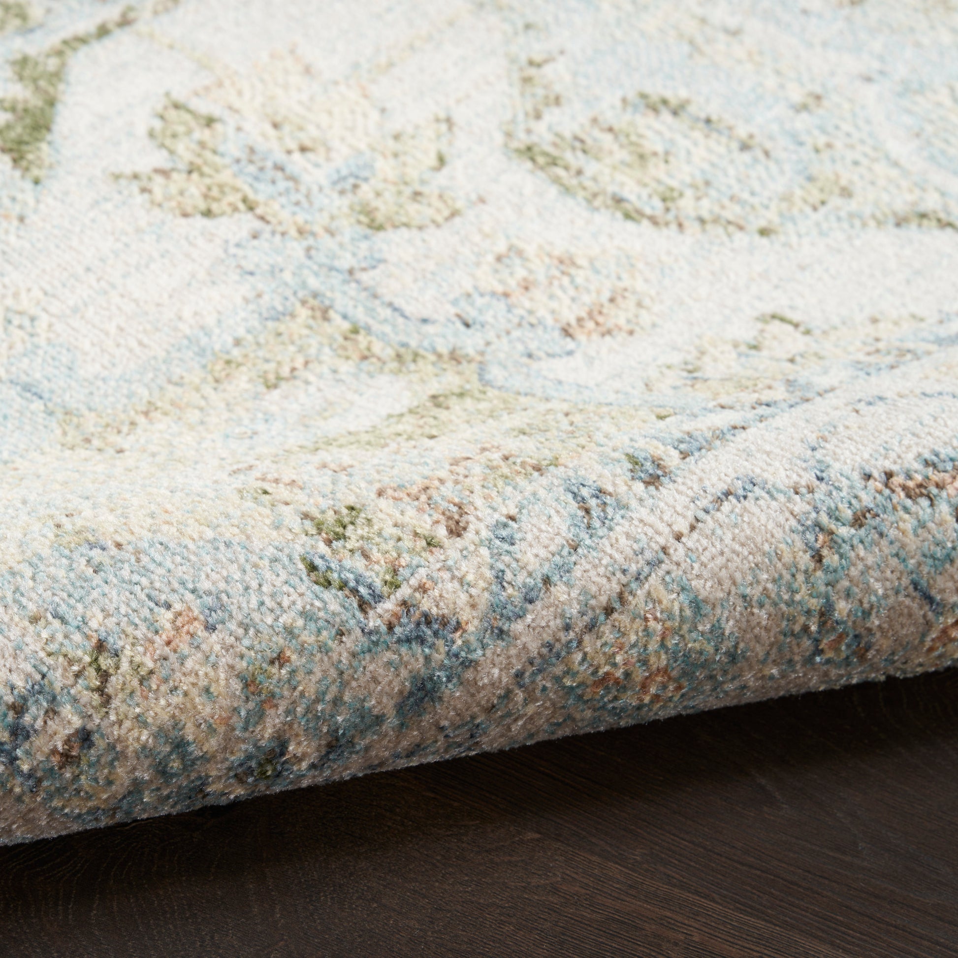 Pastel blue and cream rug with subtle floral pattern, rolled edge on dark wood floor.