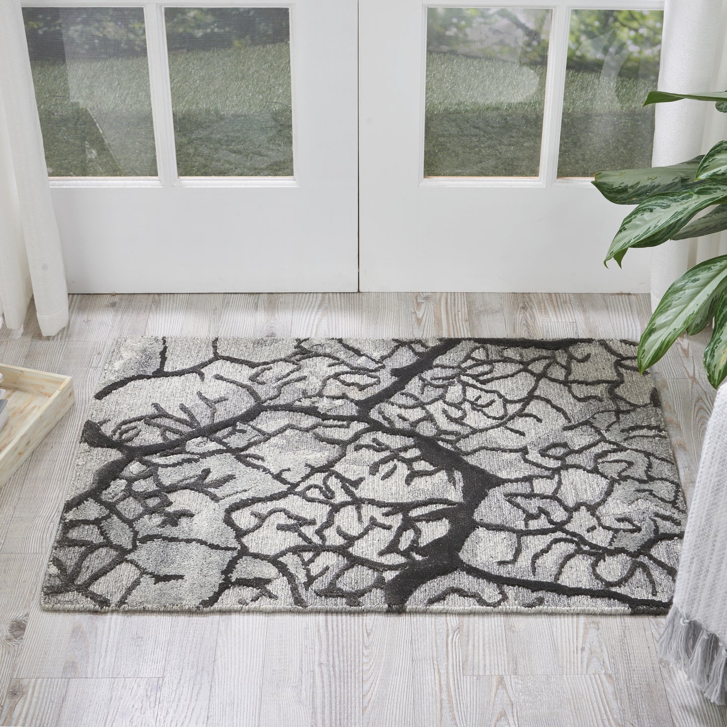 Rectangular grey rug with bold black tree-branch pattern on light wood floor near glass doors.