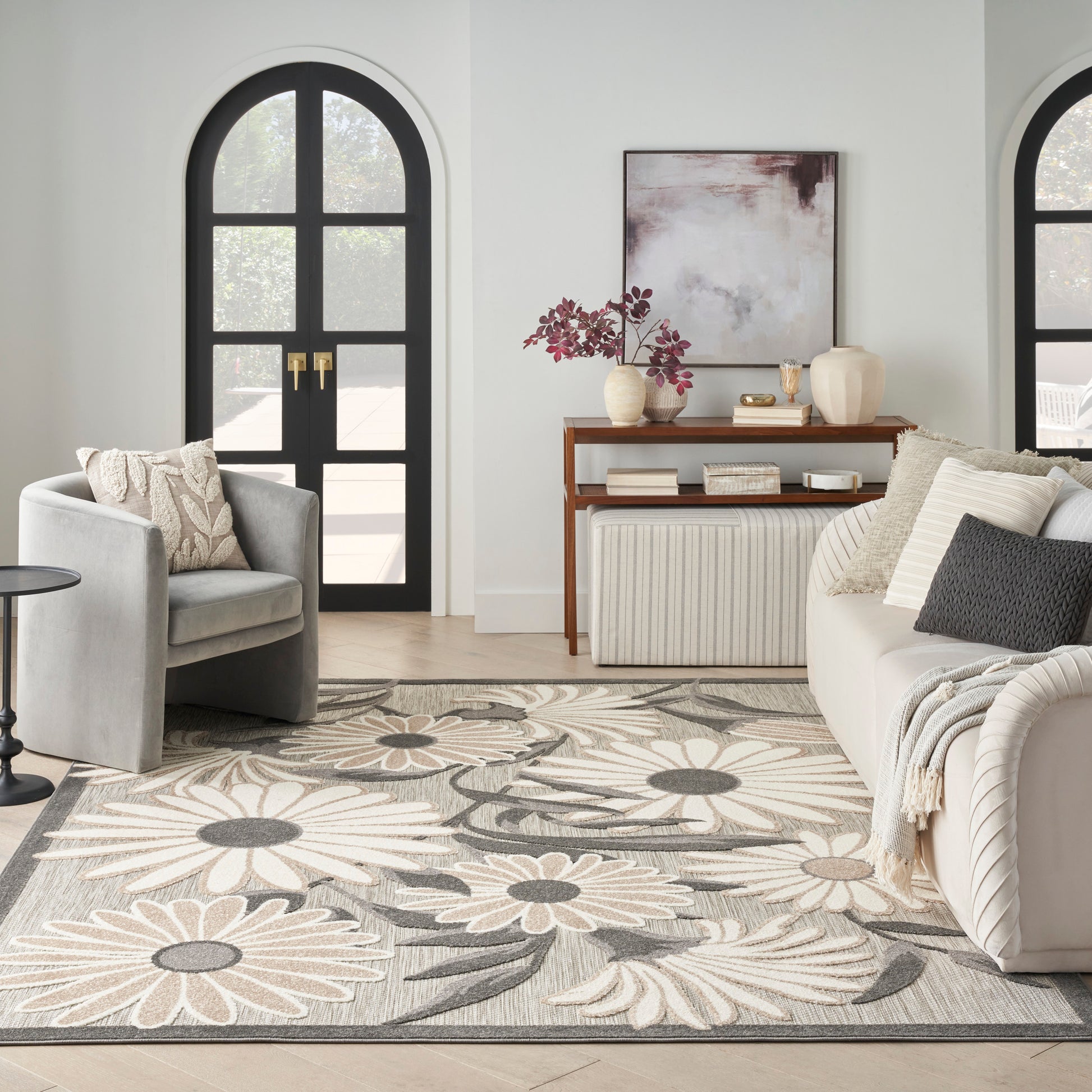 Neutral living room with a cream-gray floral rug, gray sofa, beige armchair, wooden console, and arched black doors.