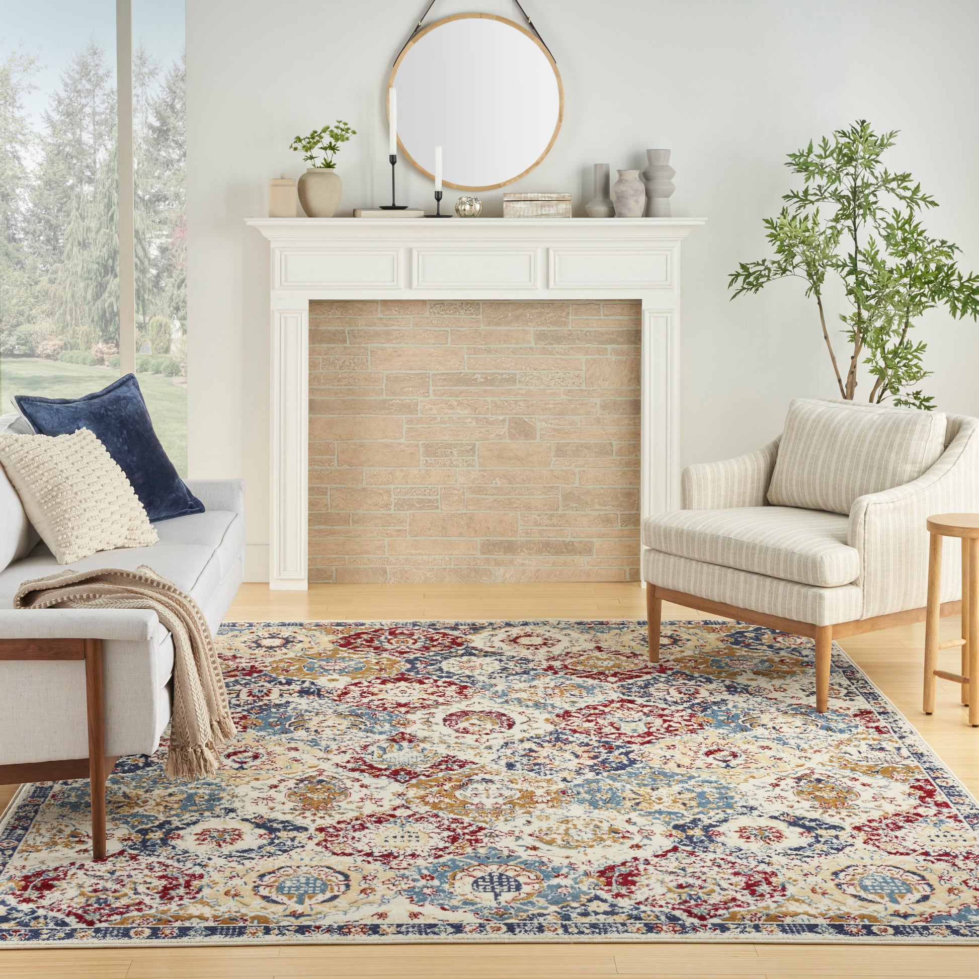 Bright living room with white mantel fireplace, round mirror, brick inset, beige armchairs, colorful Persian rug, plants.