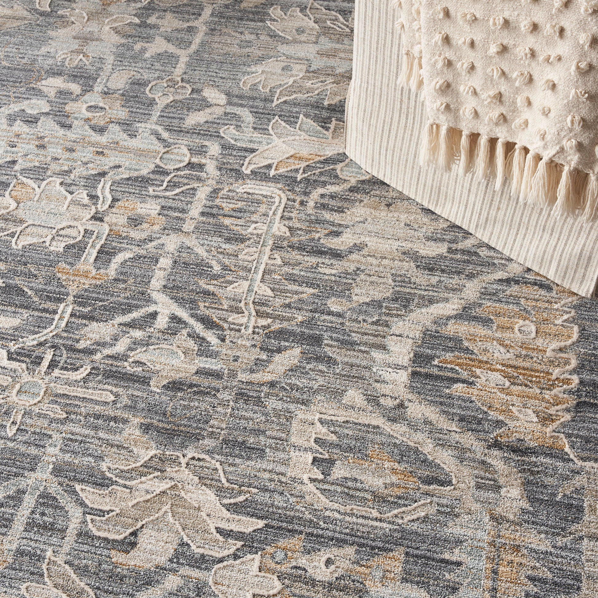 Blue-gray patterned area rug with beige floral motifs; cream textured throw with fringe at edge.