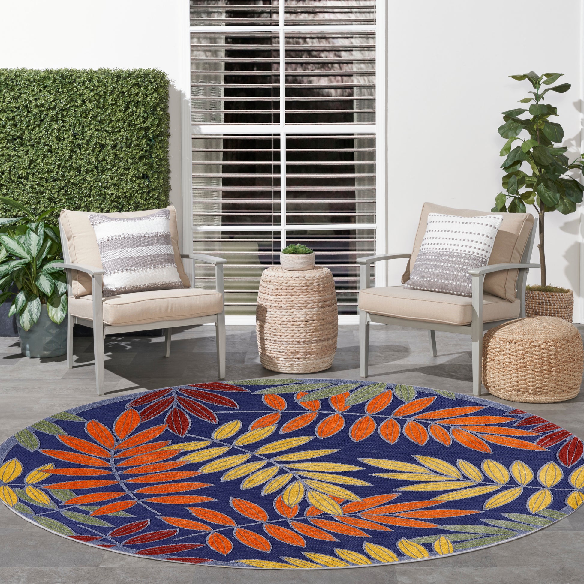 Outdoor patio with two beige cushioned chairs, round blue leaf rug in orange/yellow, wicker ottomans, plants.