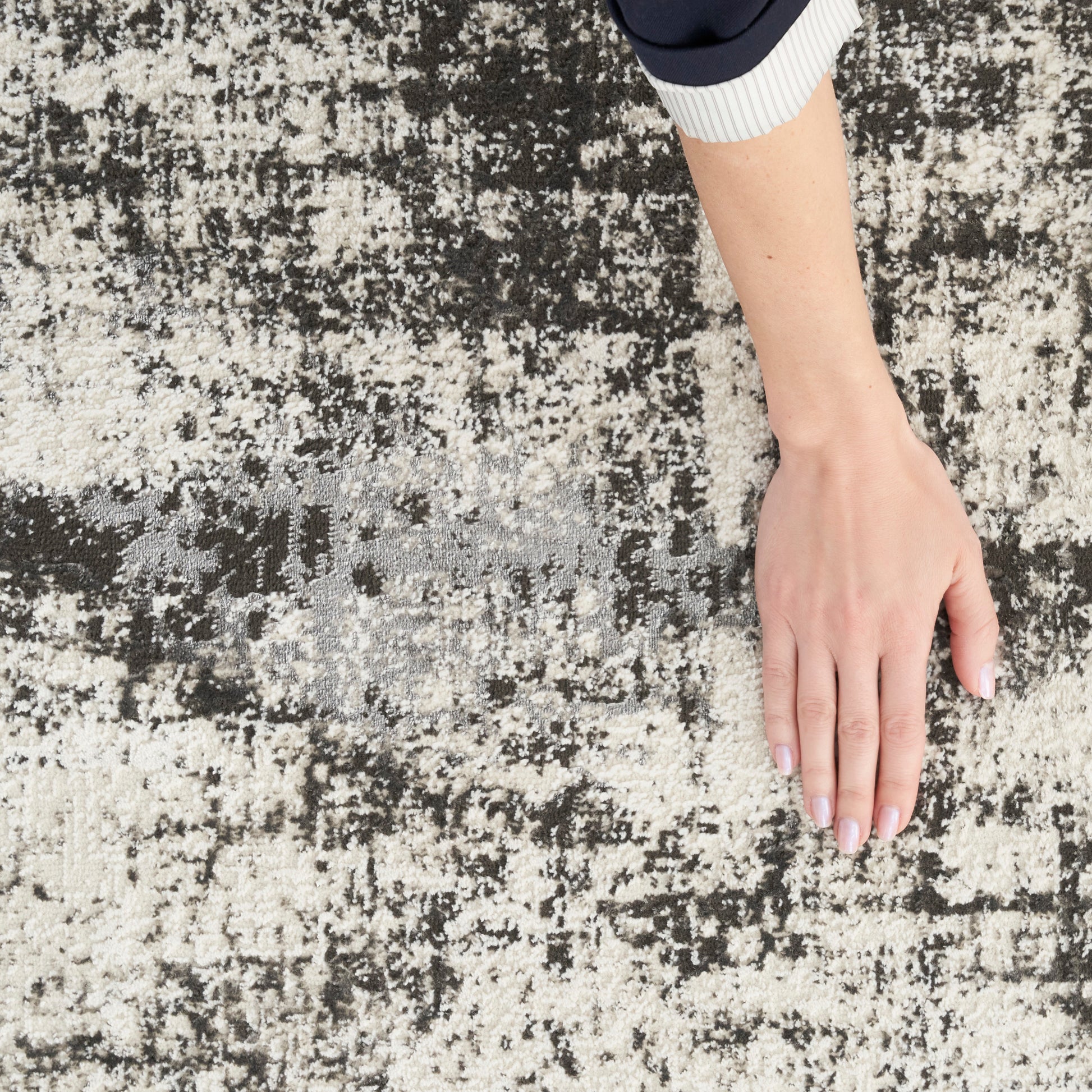 Hand with pale nails rests on beige-black abstract rug; navy and white striped sleeve cuff visible.