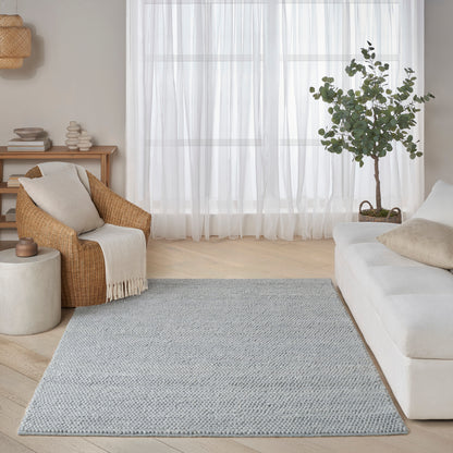 Light gray woven area rug in a serene living room with a white sofa, wicker armchair, wooden console, and potted tree.