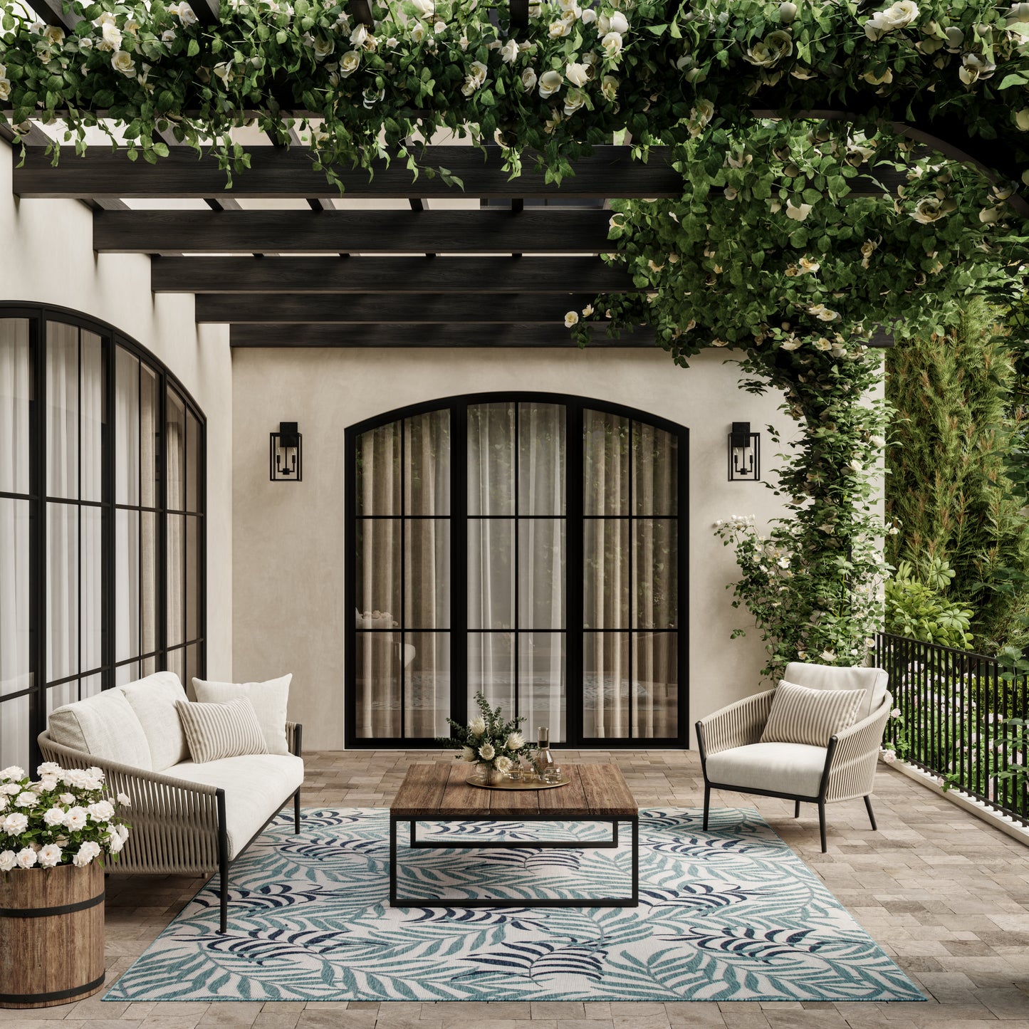 Outdoor patio with beige cushioned seating, dark metal frames, wooden coffee table, blue leaf rug, and lush greenery.