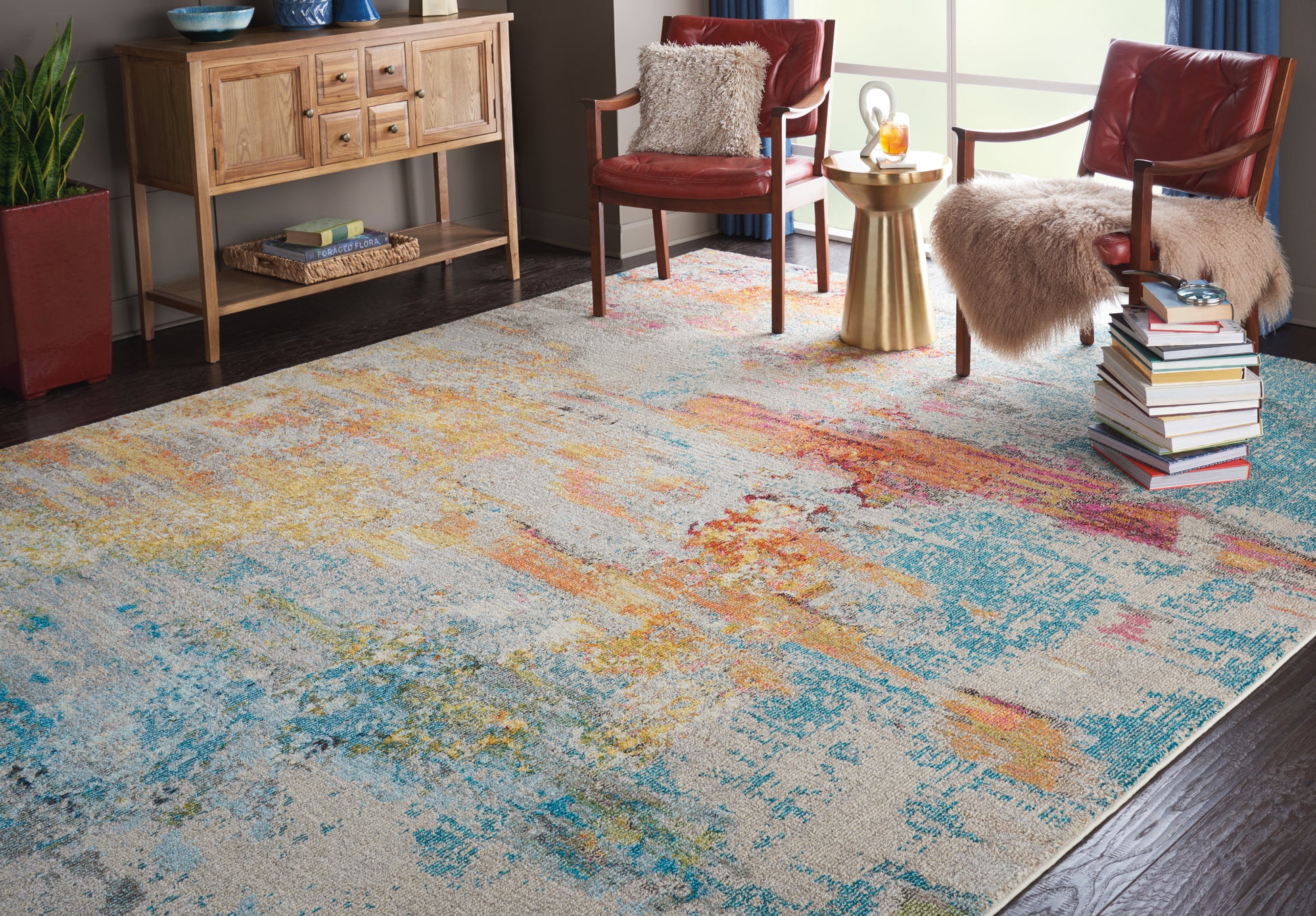 Colorful abstract area rug with blue, orange, and yellow patches in a cozy living room with red chairs, fur throws, books.