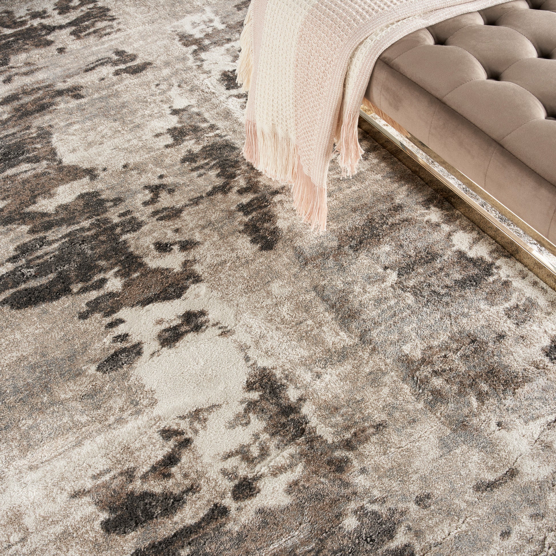 Abstract beige-taupe area rug with dark splotches; tufted beige sofa edge with a pale pink knit throw draped over.