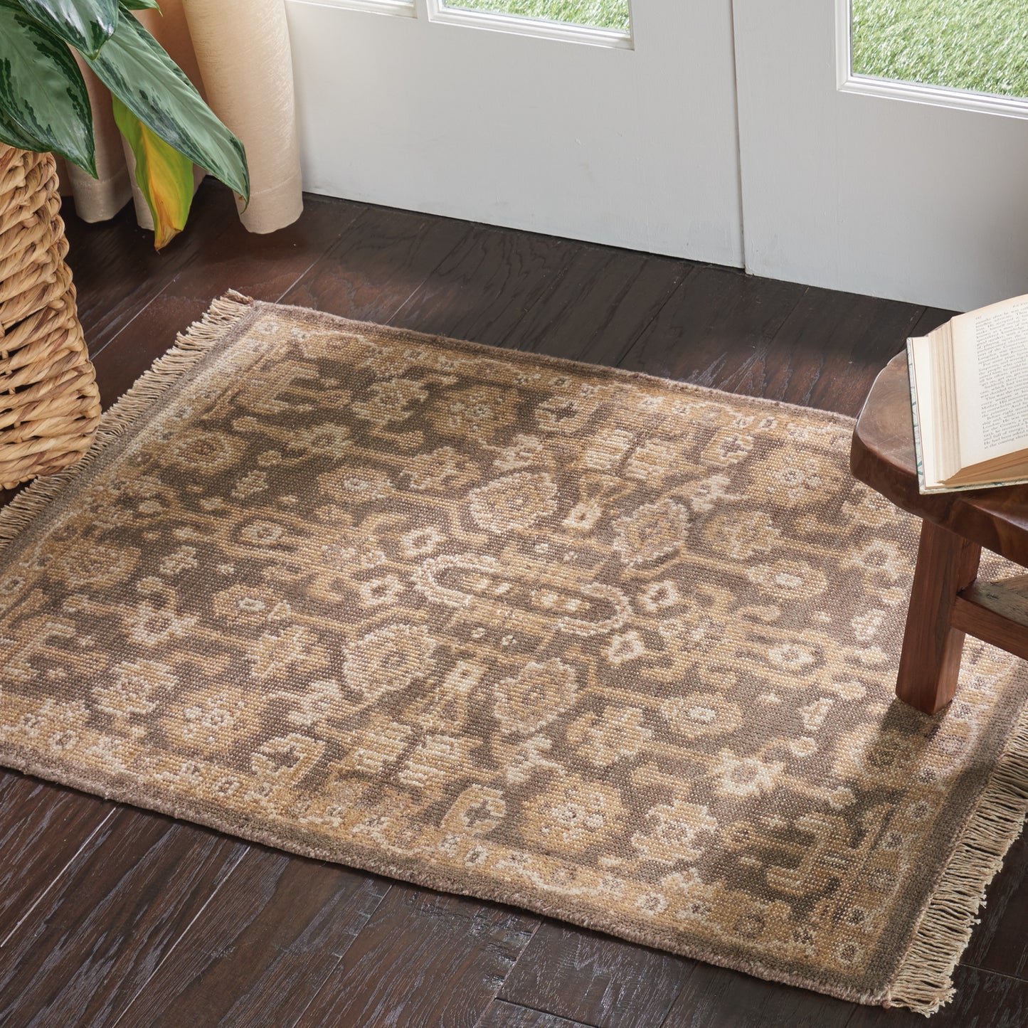 Beige-brown oriental area rug with fringe ends on dark wood floor by a door; plant basket and small wooden table with a book.