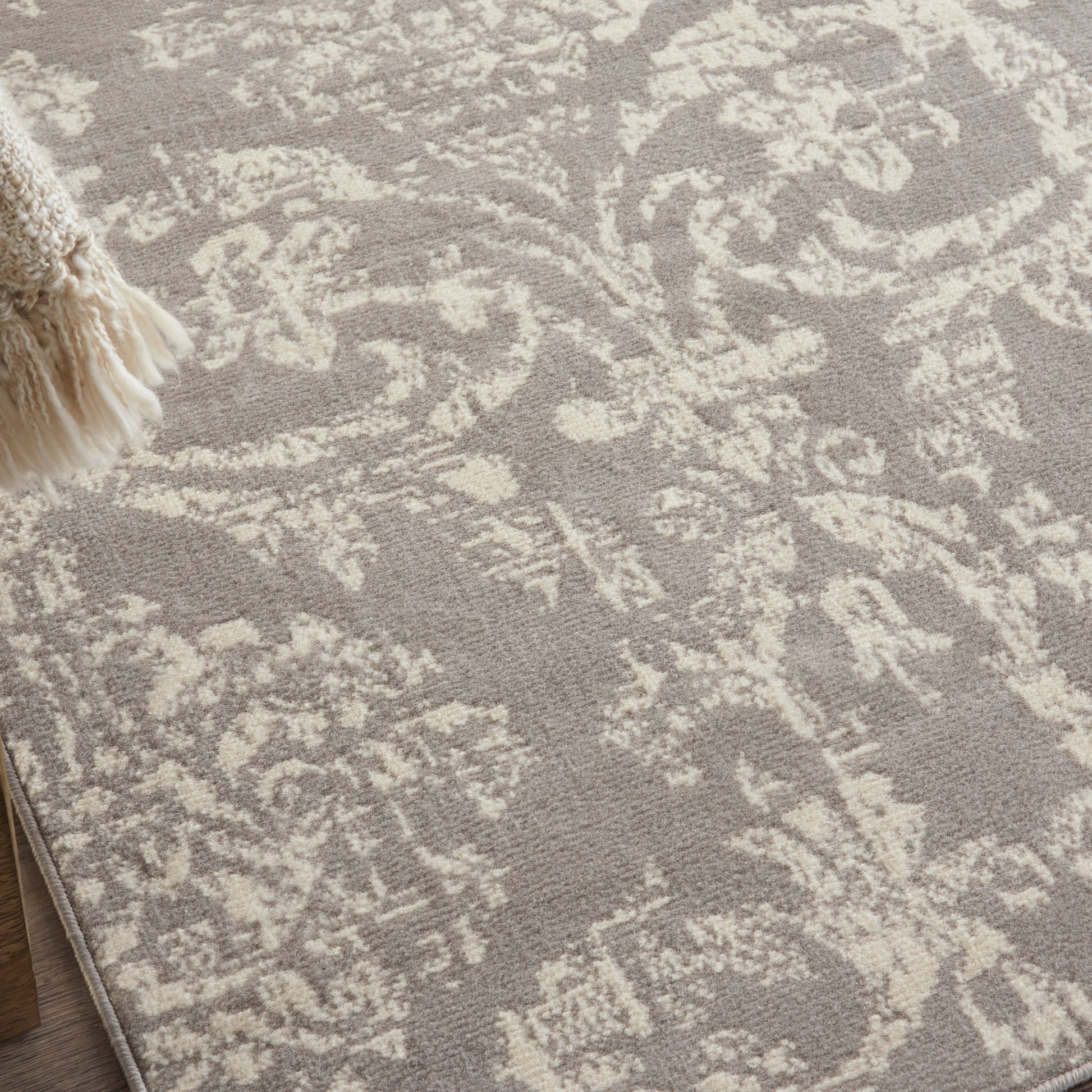 Taupe rug with cream damask floral pattern and fringe tassel trim along the edge.