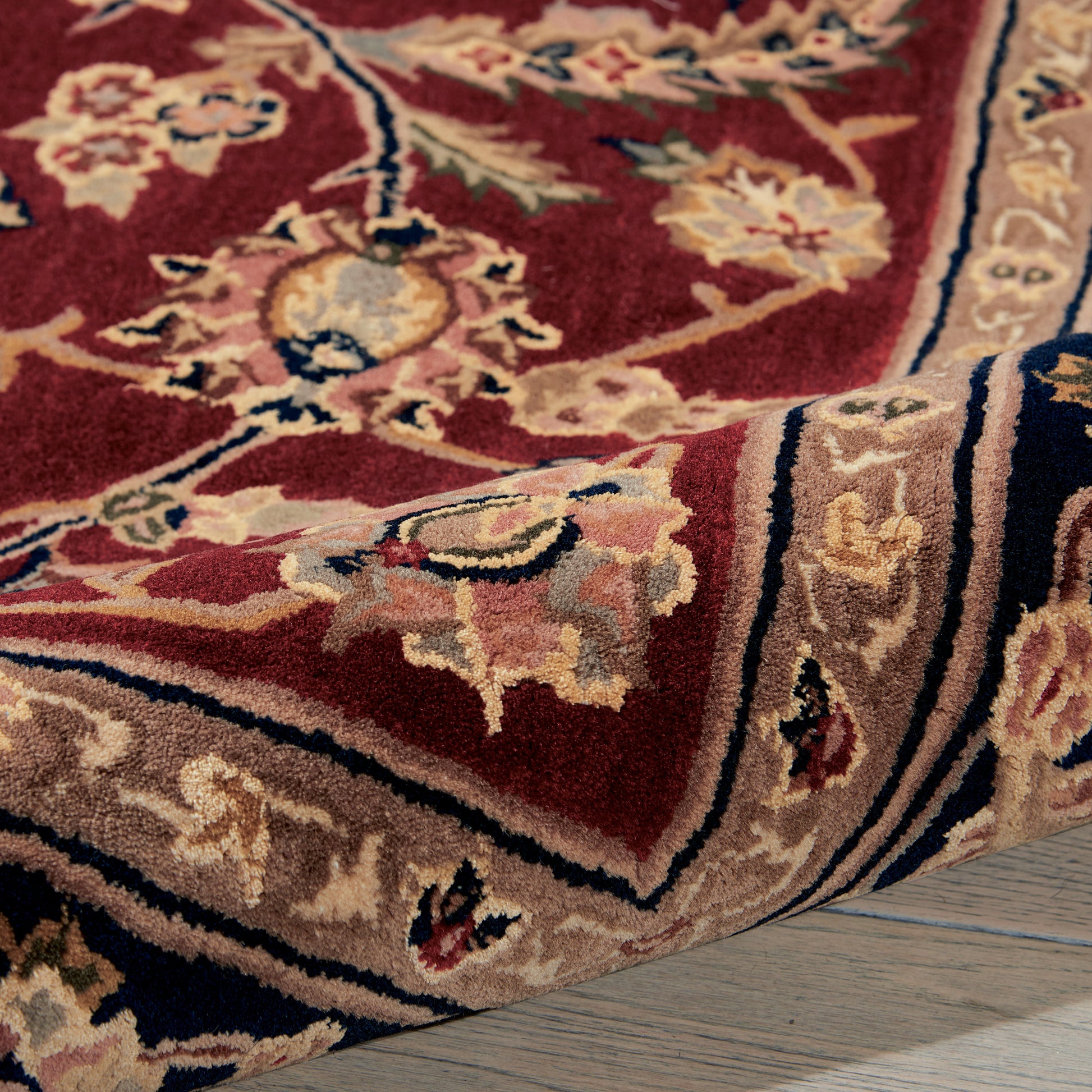 Red ornate rug with floral motifs, beige border, rolled corner, blue and ivory accents on a wooden floor.
