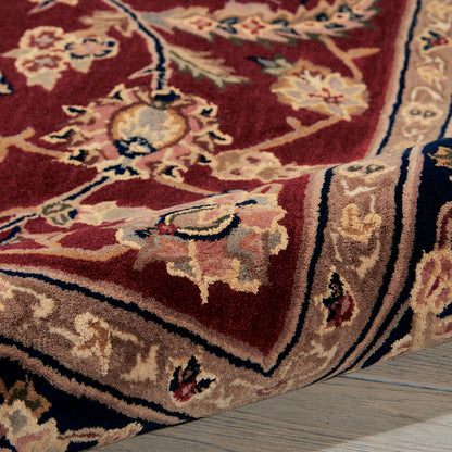 Red ornate rug with floral motifs, beige border, rolled corner, blue and ivory accents on a wooden floor.