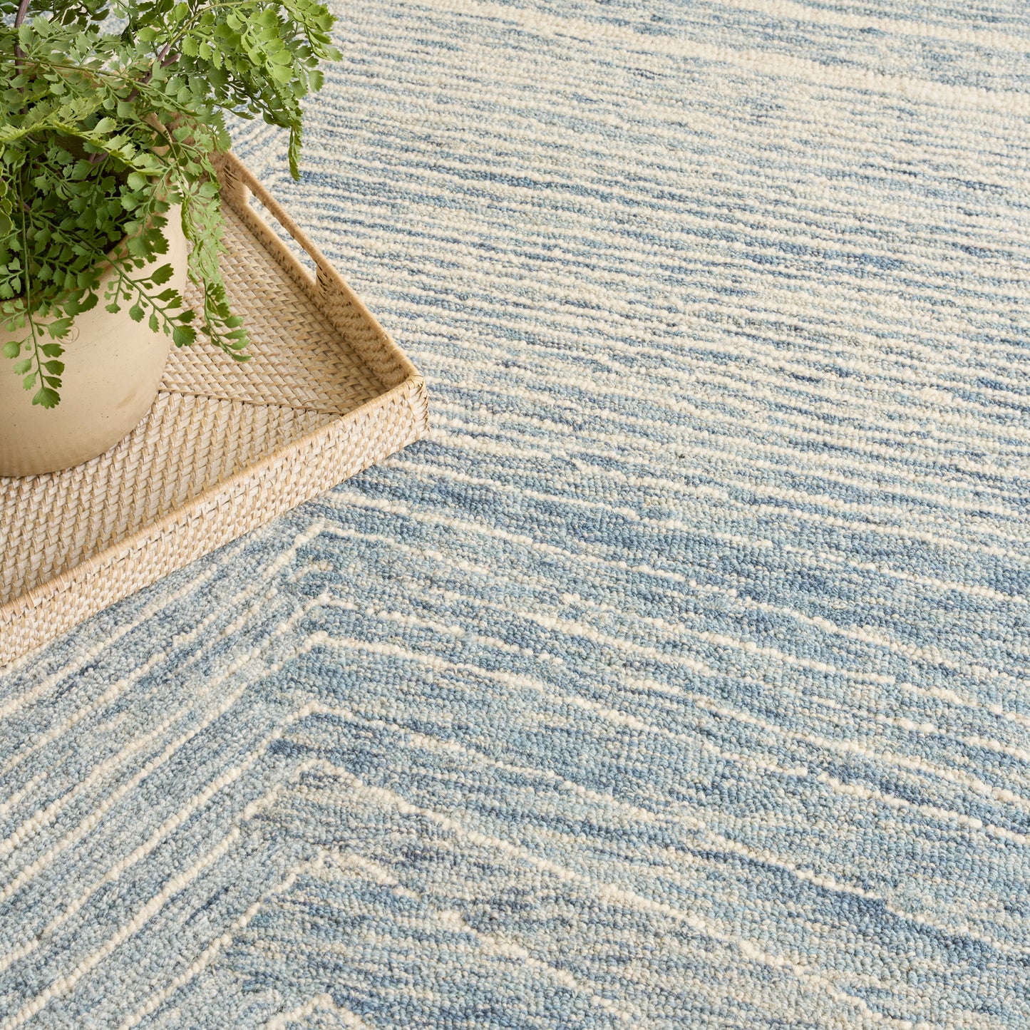 Blue and white striped textured rug with a wavy pattern, beige wicker tray and potted green plant.