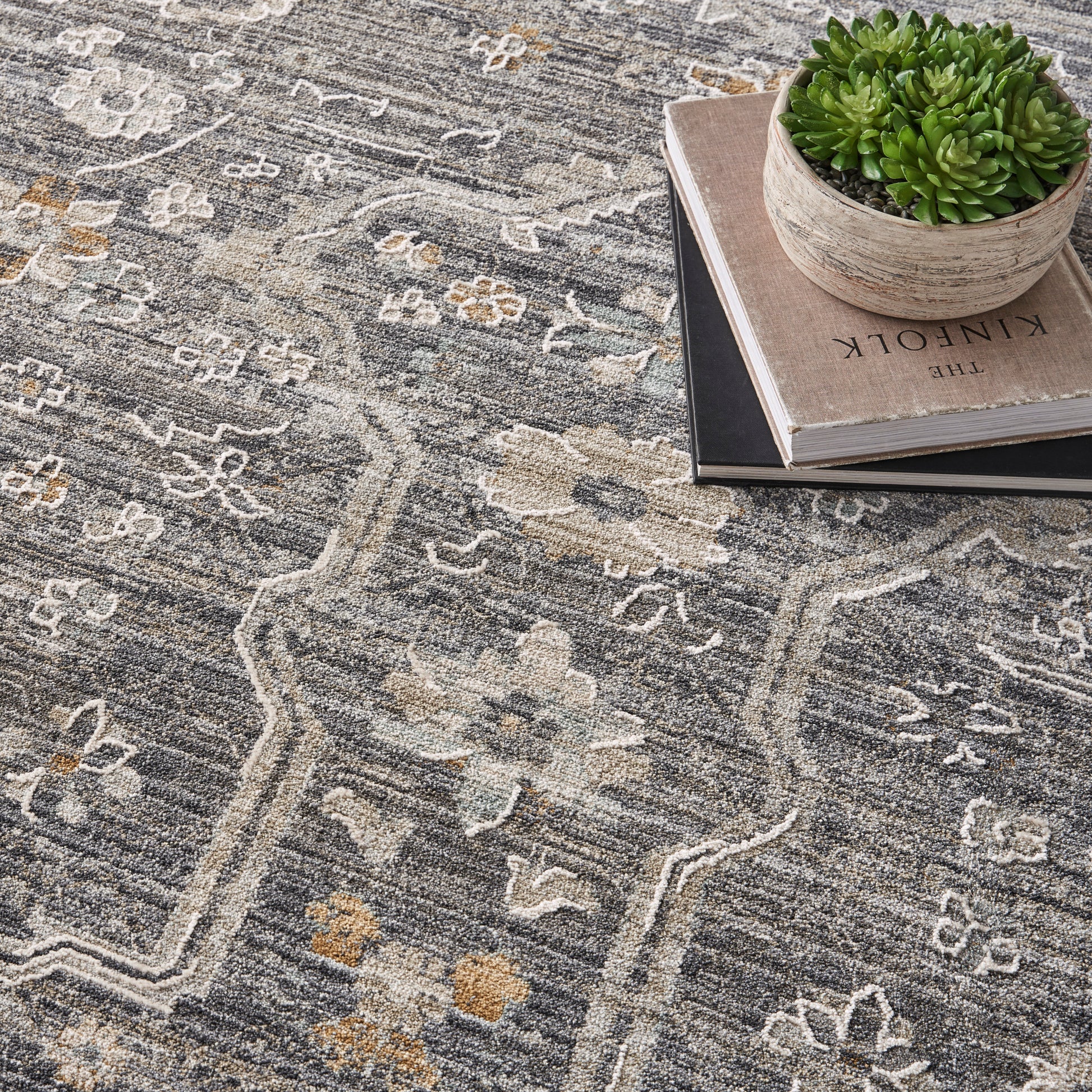 Gray oriental rug with cream floral motifs; stacked books topped by a round ceramic pot with a green succulent.