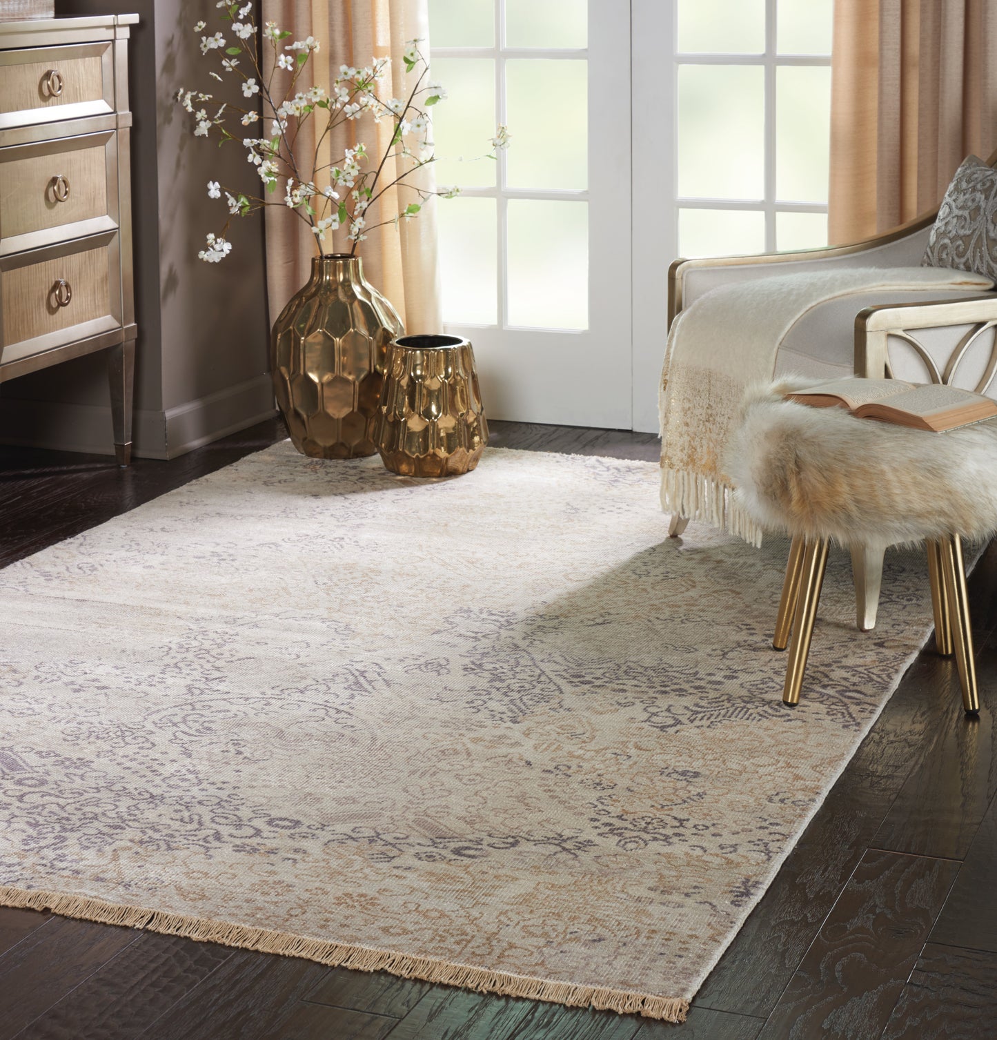 Beige damask area rug with fringe, gold hammered metal vases, fur stool with gold legs in a sunny living room.