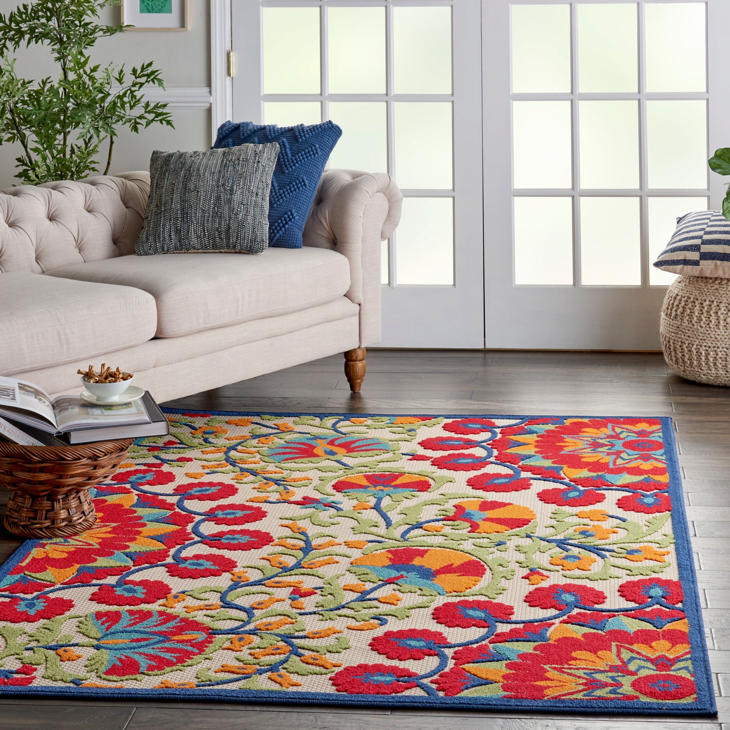 Colorful floral area rug with red, blue, yellow and orange blooms, blue border, on a wood floor beside a beige tufted sofa.