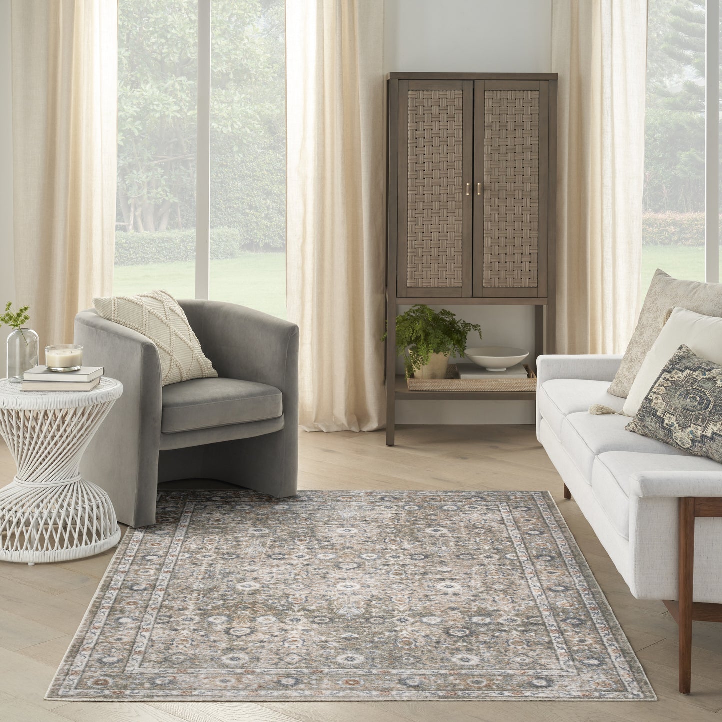 Cozy living room with gray armchairs, white sofa, beige ornate area rug, woven cabinet, white lattice side table, greenery.