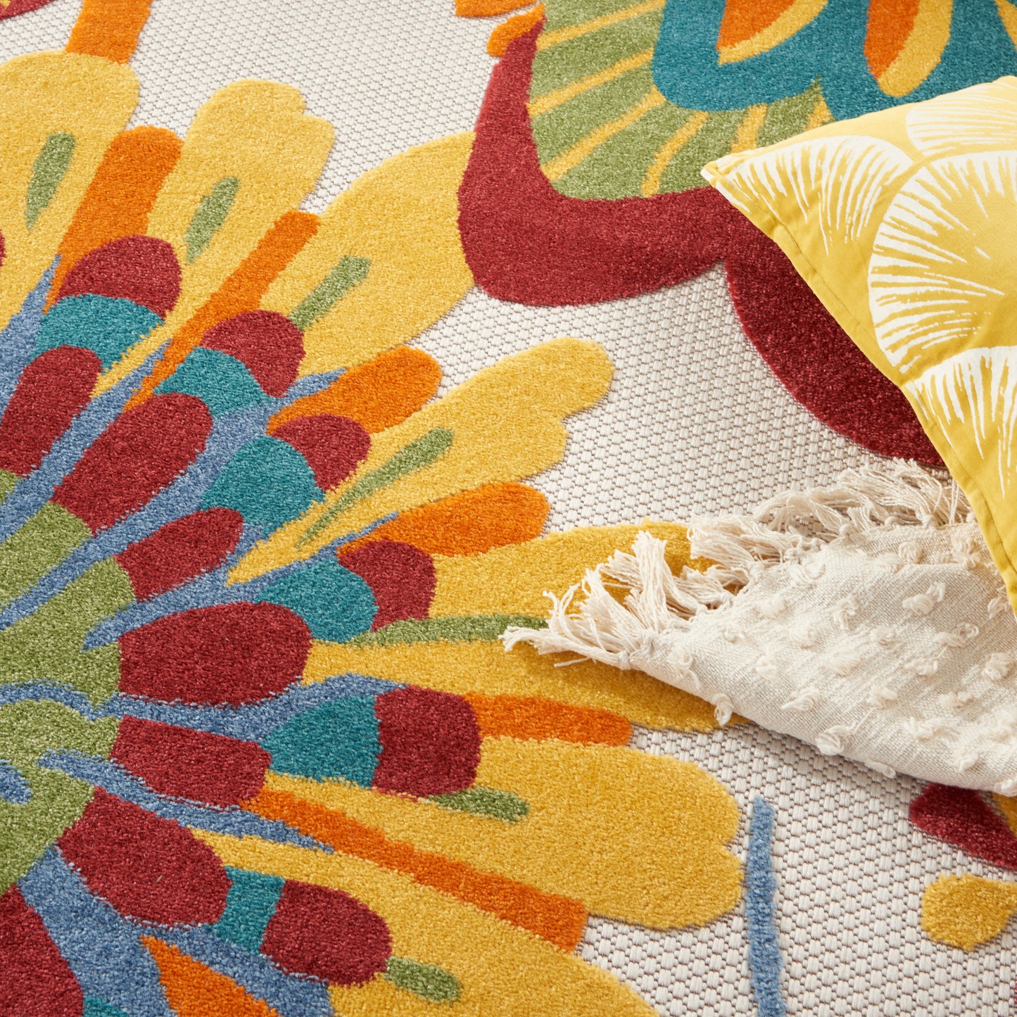 Bright multicolor round floral rug with yellow, teal, orange petals and fringe, alongside a yellow leaf-patterned pillow.