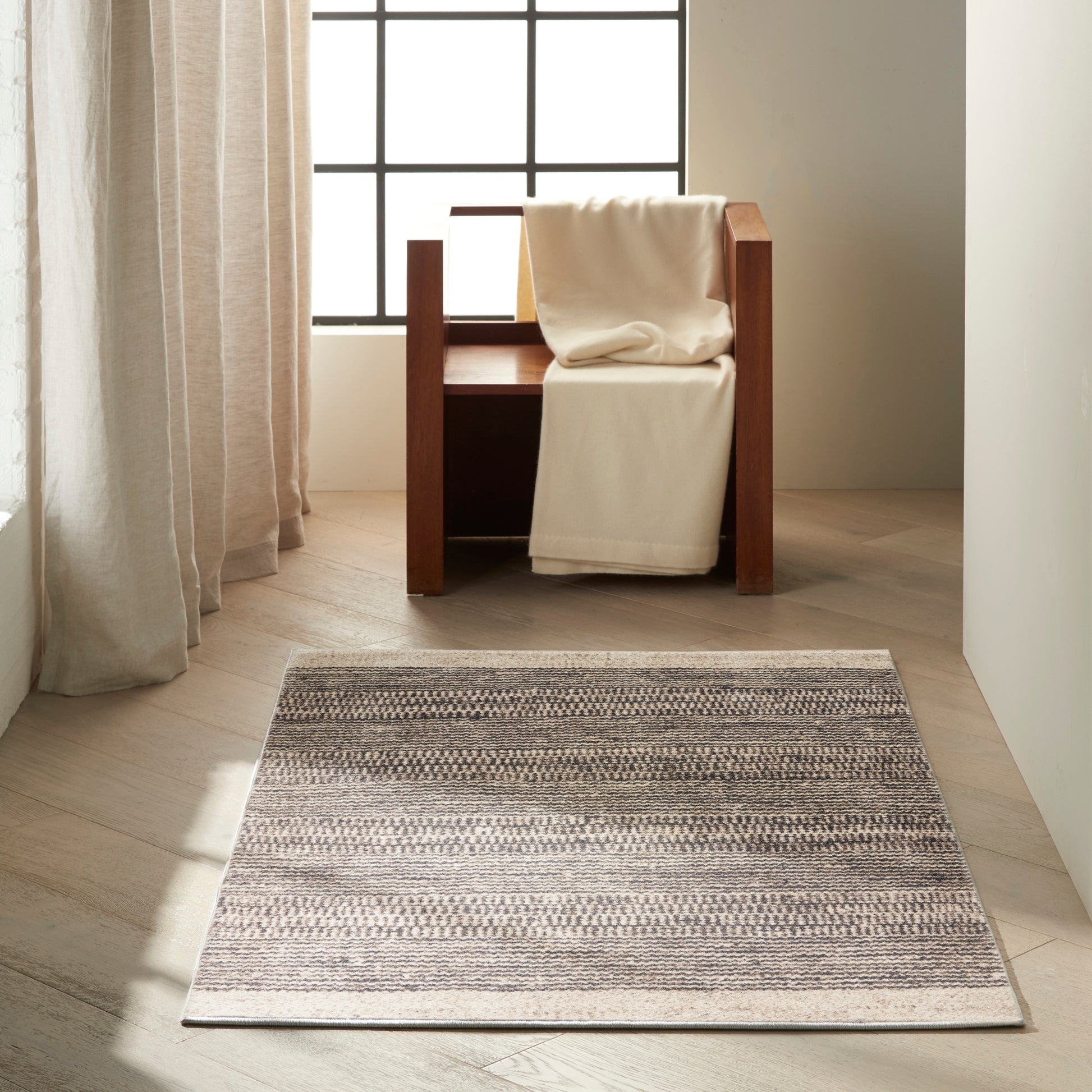 Wooden-framed armchair with a cream blanket draped over it, beside a gray-beige striped rug in a sunlit minimalist room.