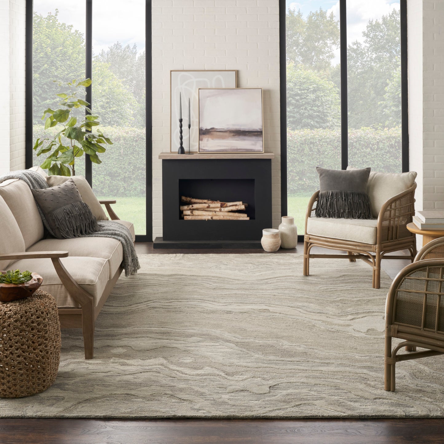 Cozy contemporary lounge with beige sofa, rattan armchairs, neutral rug, fireplace, potted plant, and art by large windows.