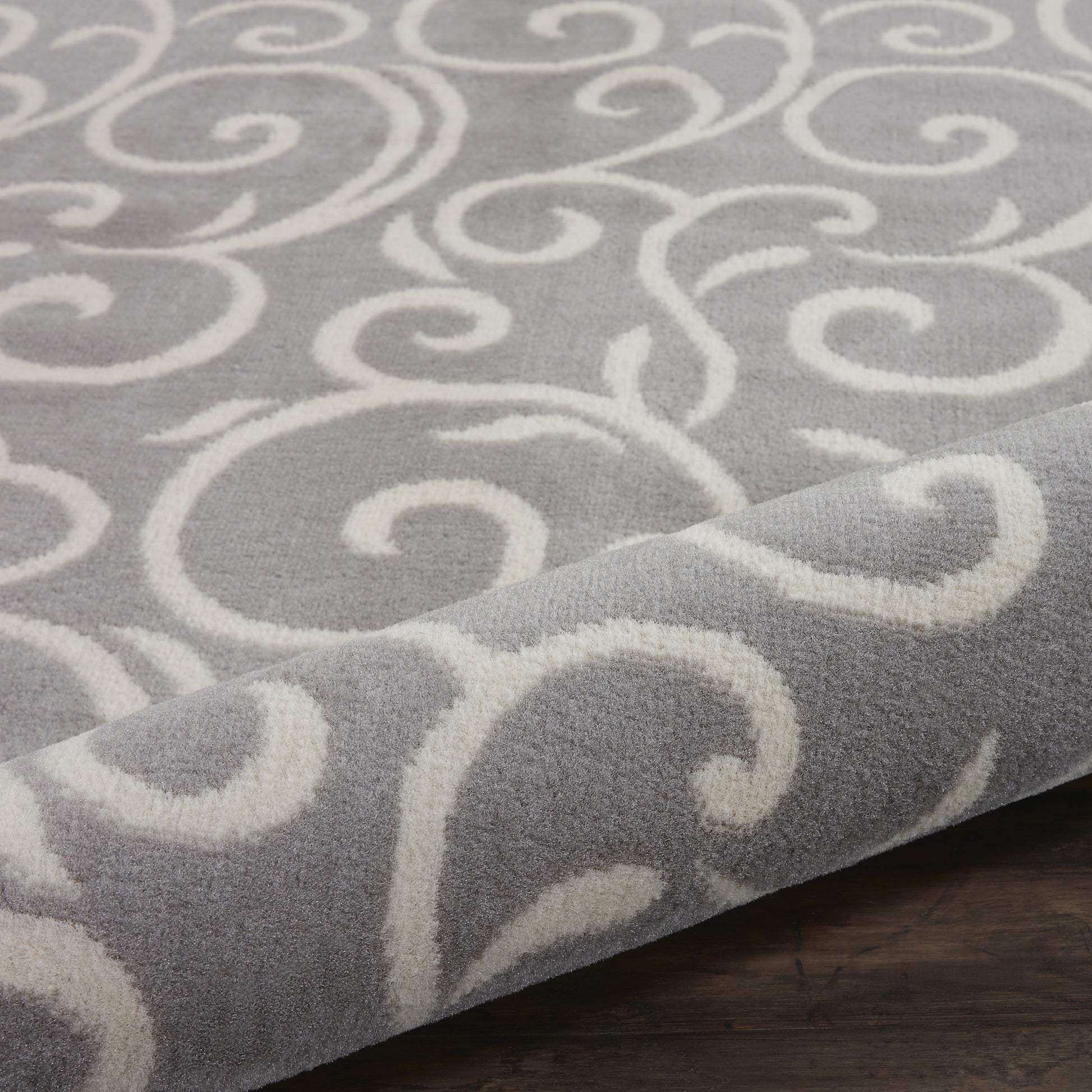 Gray rug with white swirling scroll pattern, soft plush texture, rolled corner on dark wooden floor.