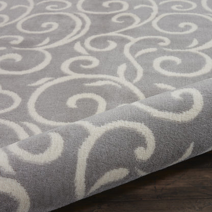 Gray rug with white swirling scroll pattern, soft plush texture, rolled corner on dark wooden floor.
