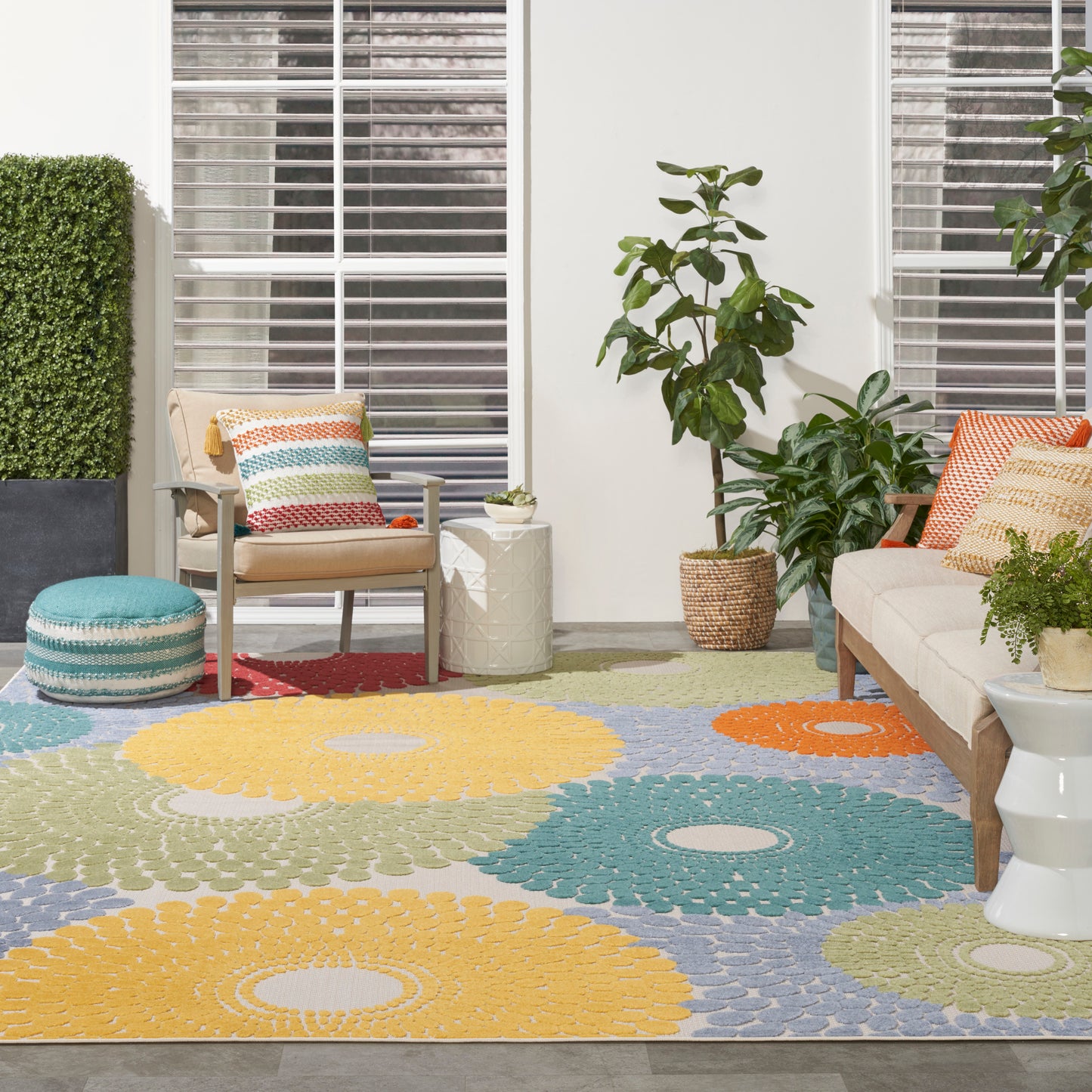 Outdoor patio with colorful circular-pattern rug, beige sofa and chair, potted plants, white side tables, vibrant cushions.