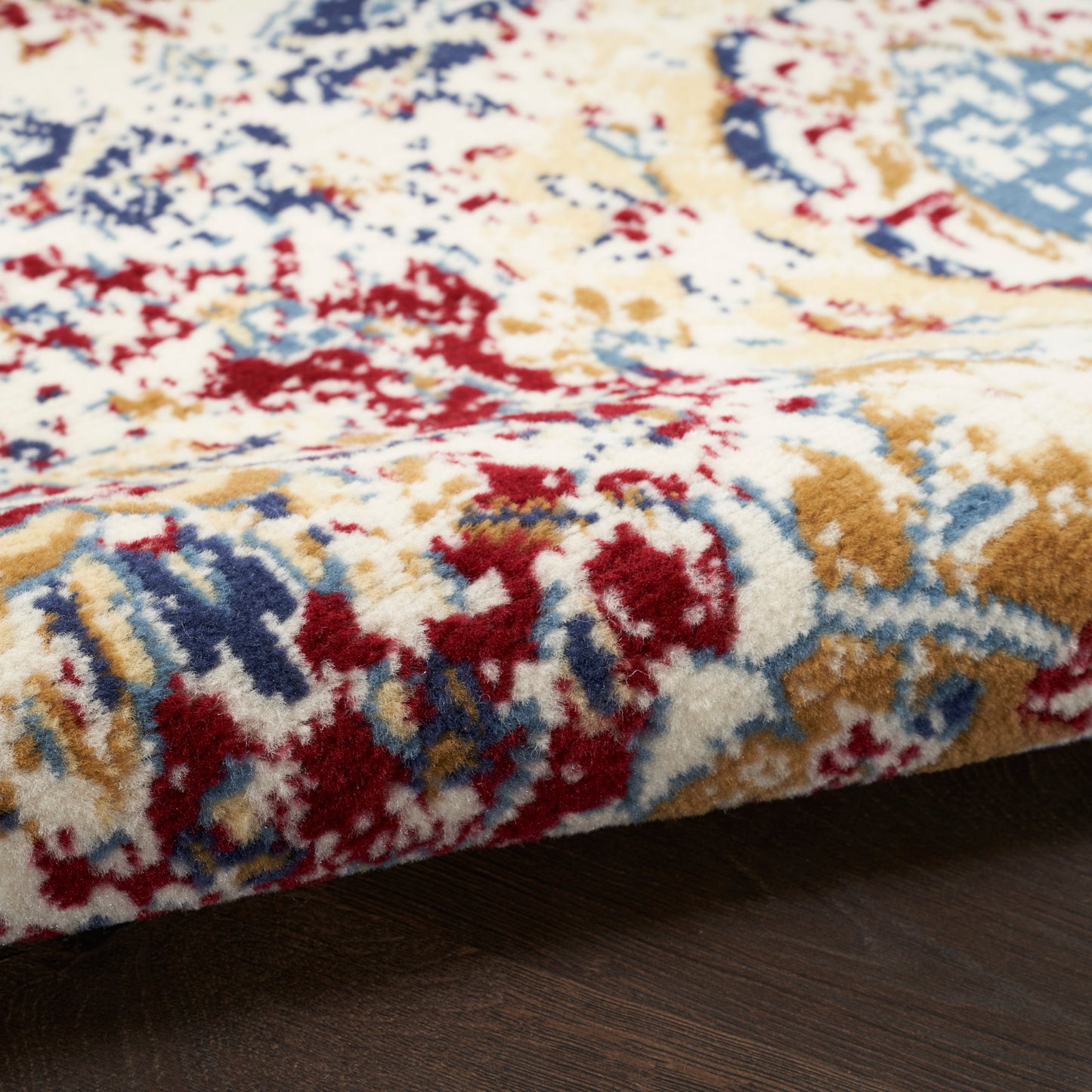 Plush area rug in red, blue, ivory and gold with a traditional pattern, on dark wood floor.