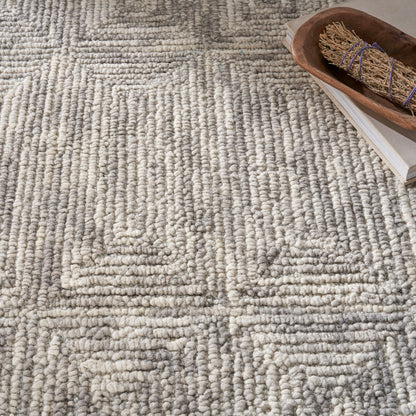 Beige-gray looped pile rug with geometric square patterns; wooden bowl with a bundled sage stick sits atop a book.