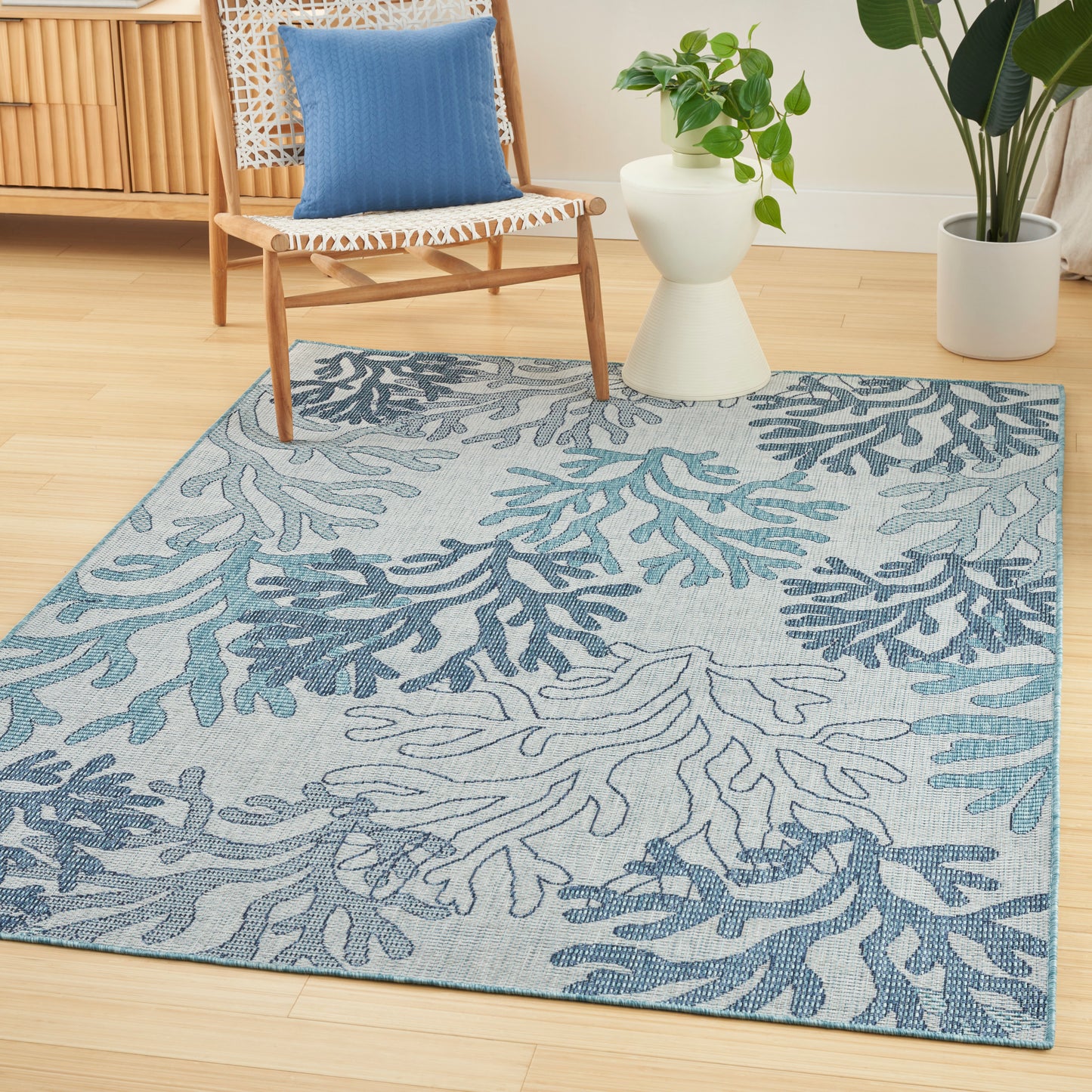 Blue-toned area rug with teal coral pattern in a bright living room, beside a woven chair and green plants.