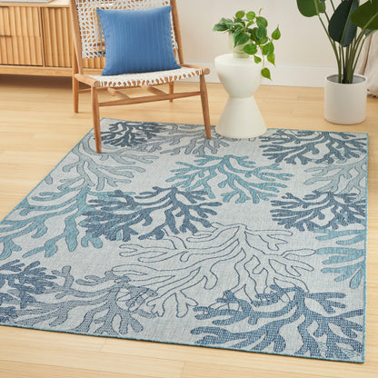 Blue-toned area rug with teal coral pattern in a bright living room, beside a woven chair and green plants.