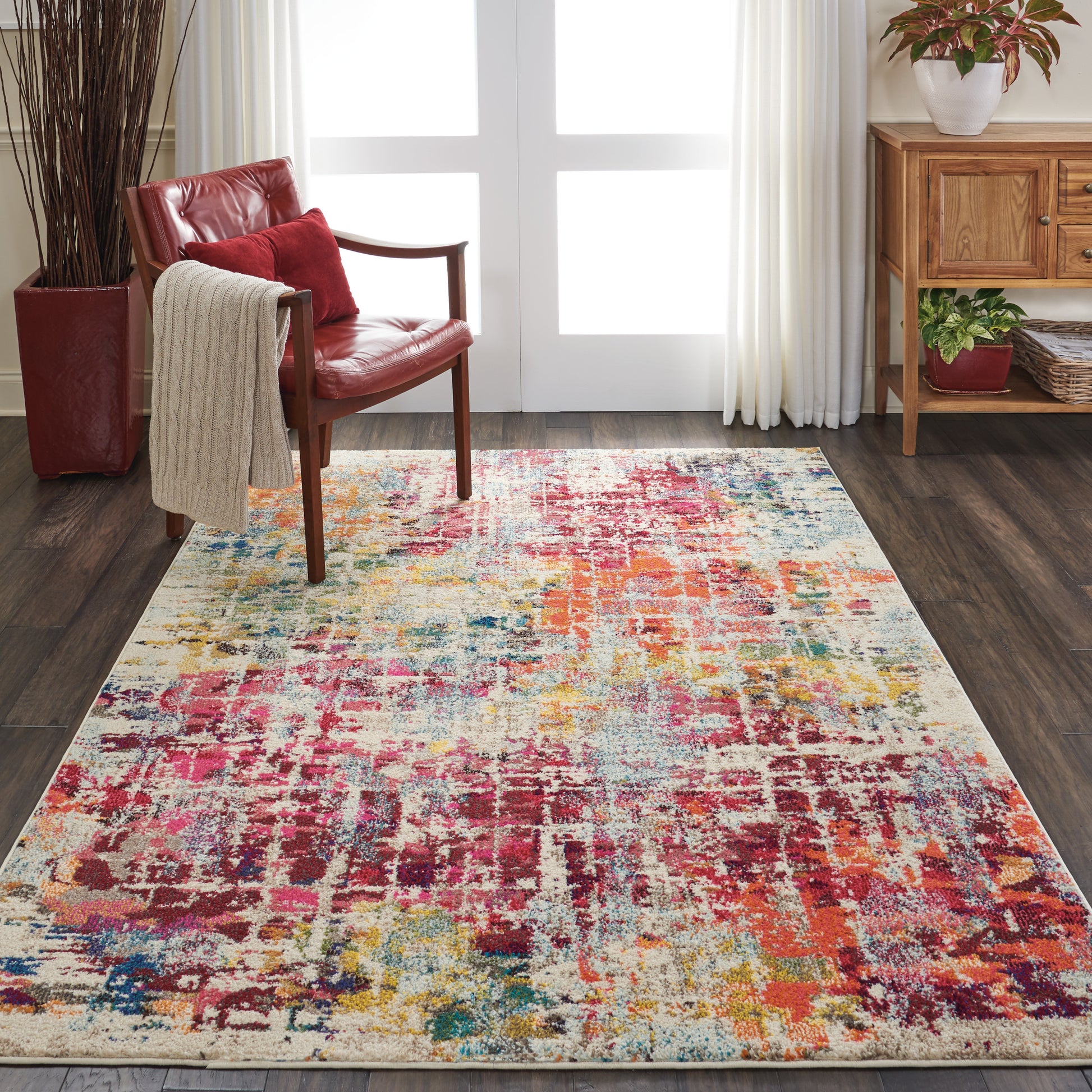 Colorful abstract area rug on dark wood floor beside red leather chair with beige throw, wooden sideboard and potted plants.