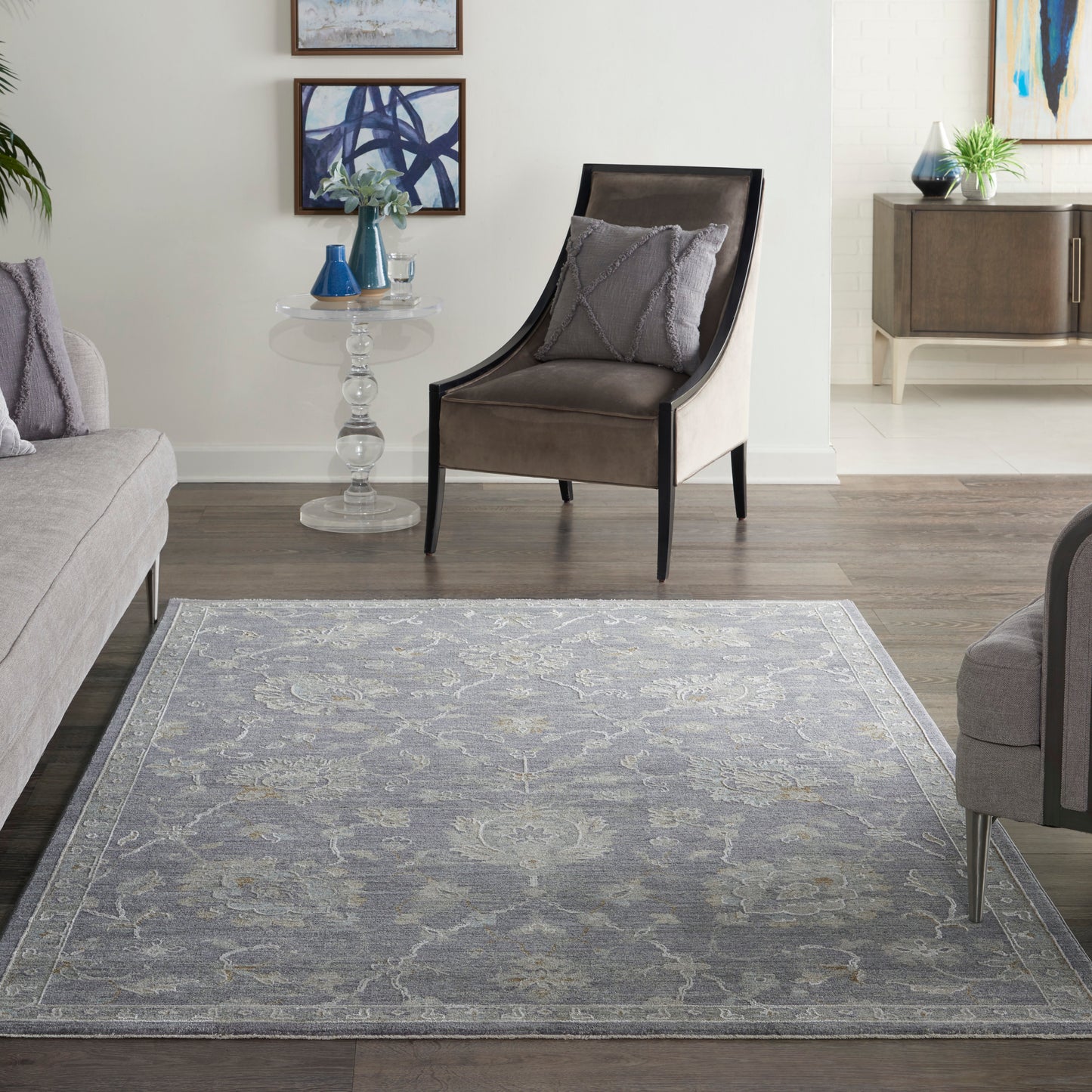 Gray-blue ornate area rug anchors a modern living room with a curved-back accent chair, glass side table, and abstract art.