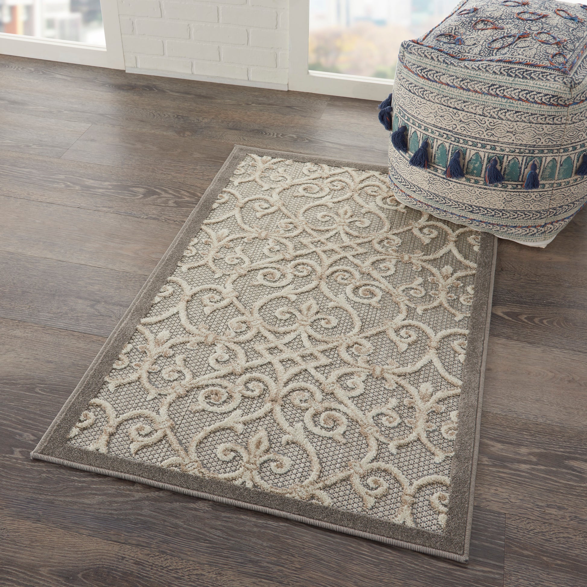 Gray and beige ornate area rug with scrolling pattern on dark wood floor near a window; round blue-tassel pouf nearby.