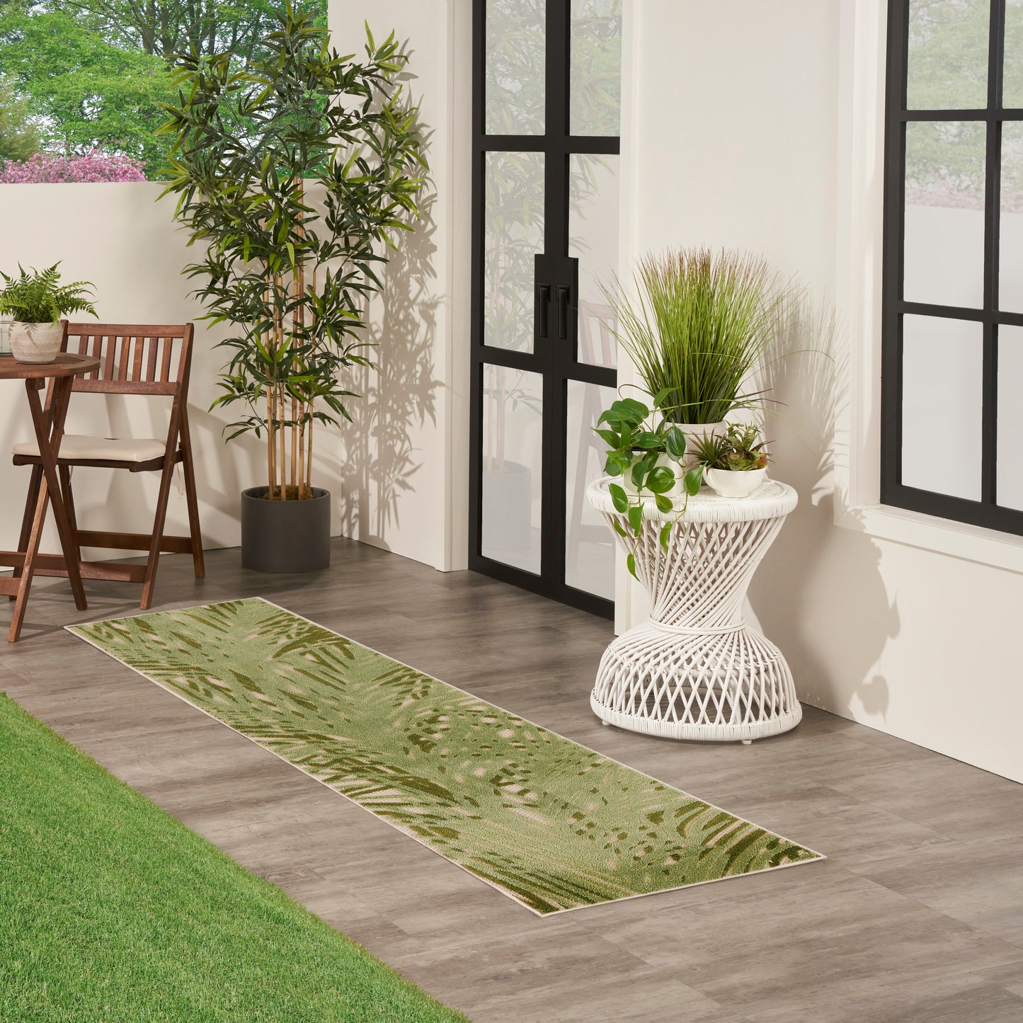 Leaf-patterned green runner rug on wood floor beside glass-framed doors, with potted plants & white wicker plant stand.
