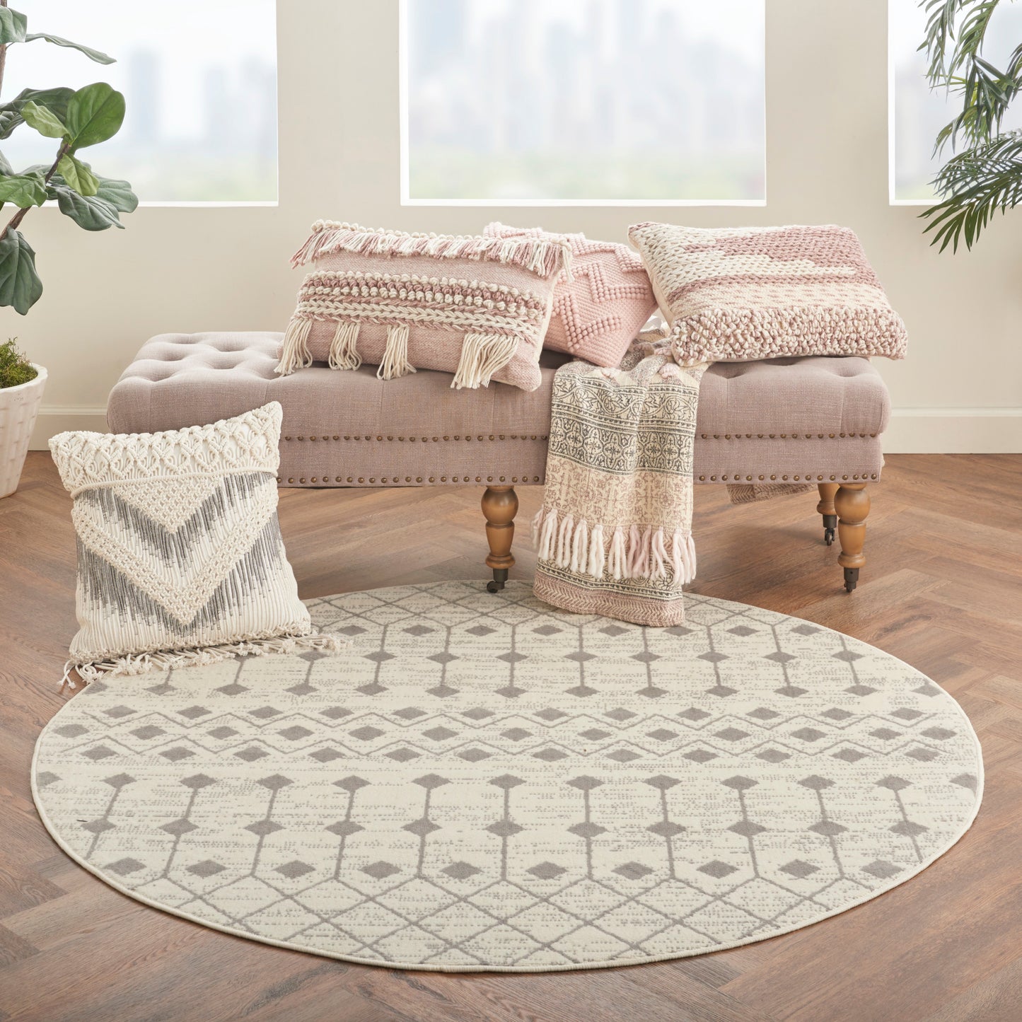 Boho round beige/cream rug with geometric lattice pattern on wooden floor, beside pink tufted ottoman and fringed pillows.