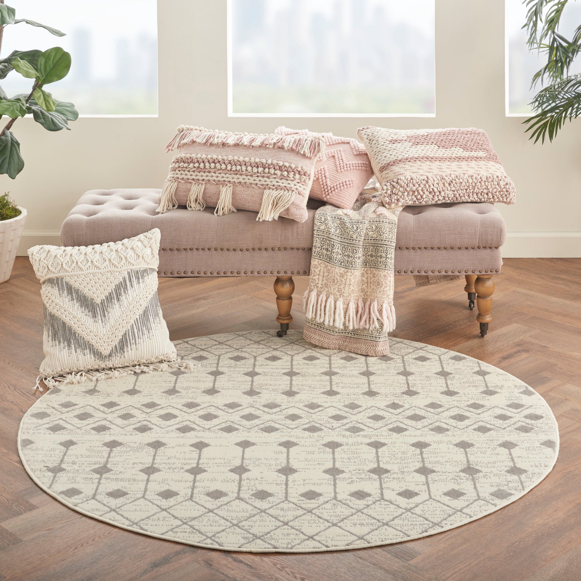 Boho round beige/cream rug with geometric lattice pattern on wooden floor, beside pink tufted ottoman and fringed pillows.