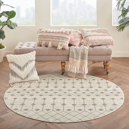 Boho round beige/cream rug with geometric lattice pattern on wooden floor, beside pink tufted ottoman and fringed pillows.