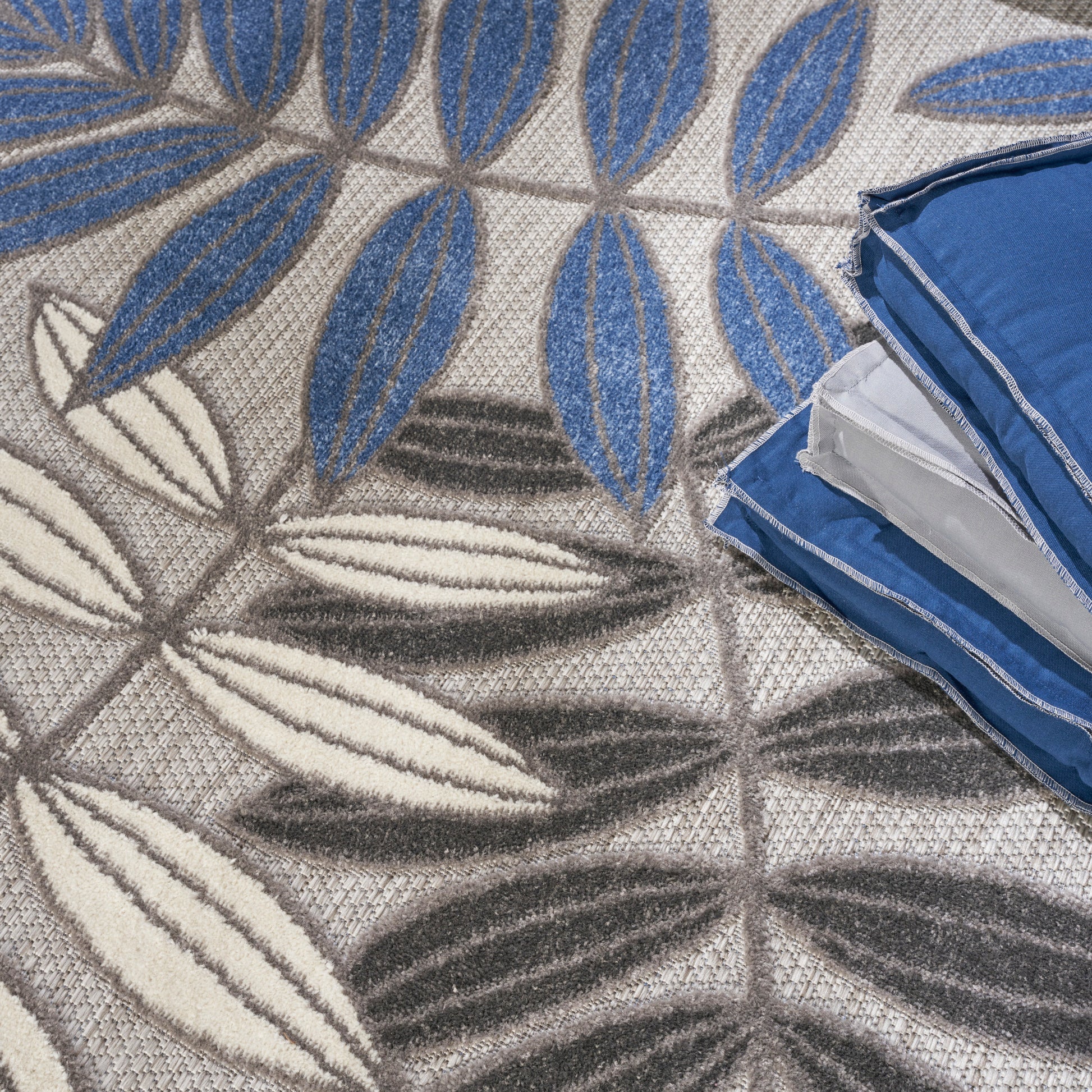 Folded blue bedding with white piping on a beige rug featuring large blue leaf motifs.