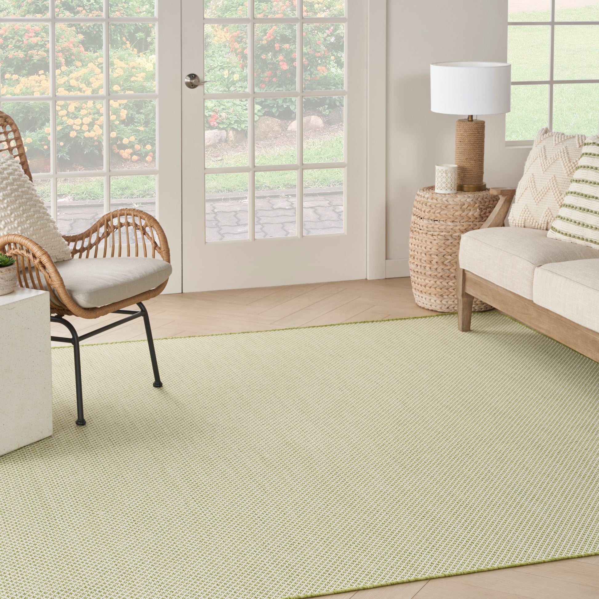 Bright living room with pale yellow-green woven rug, rattan chair, beige sofa, wicker side table with lamp, and French doors.