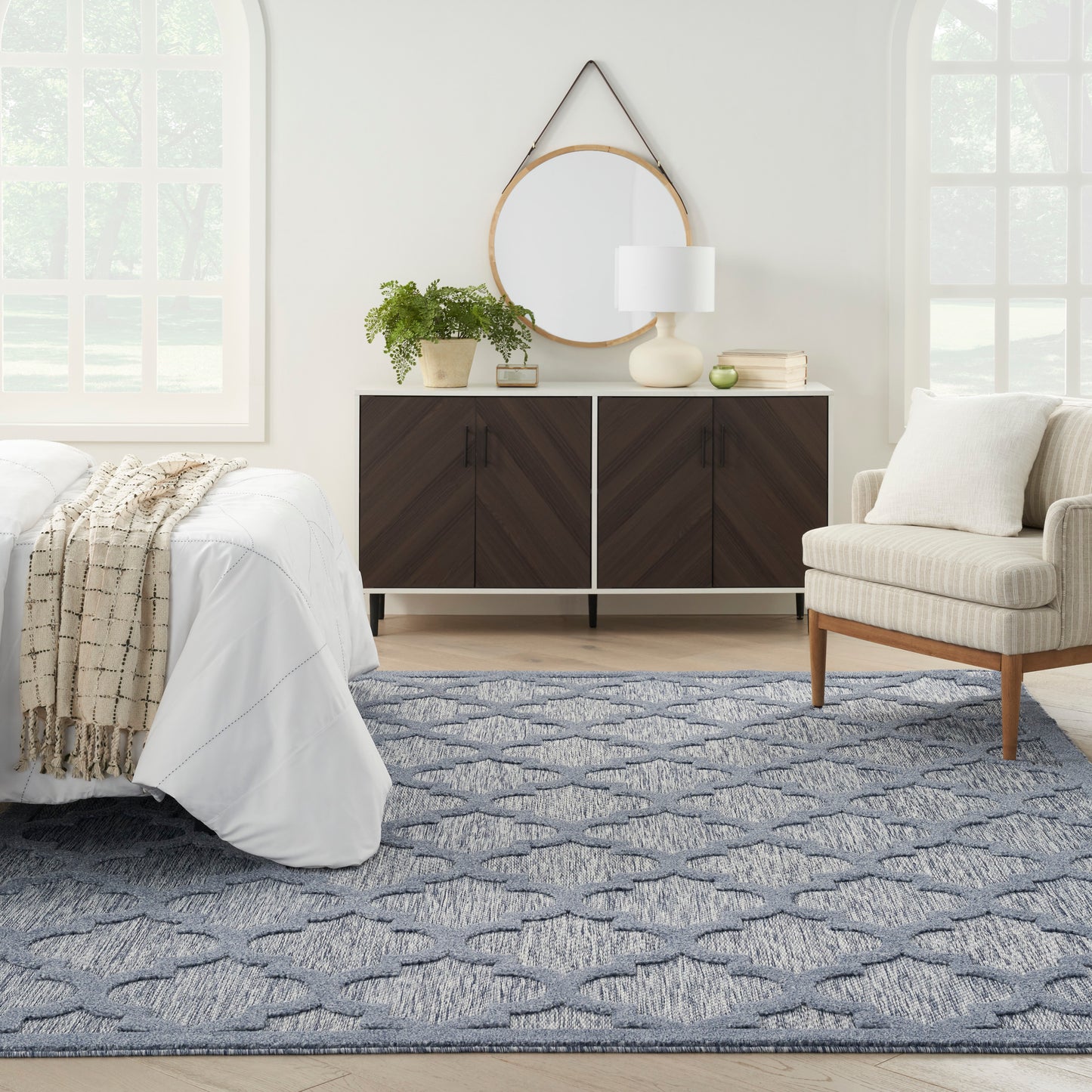 Blue rug under dark wood cabinet, round mirror, white lamp, plant, beige lounge chair in bright bedroom.