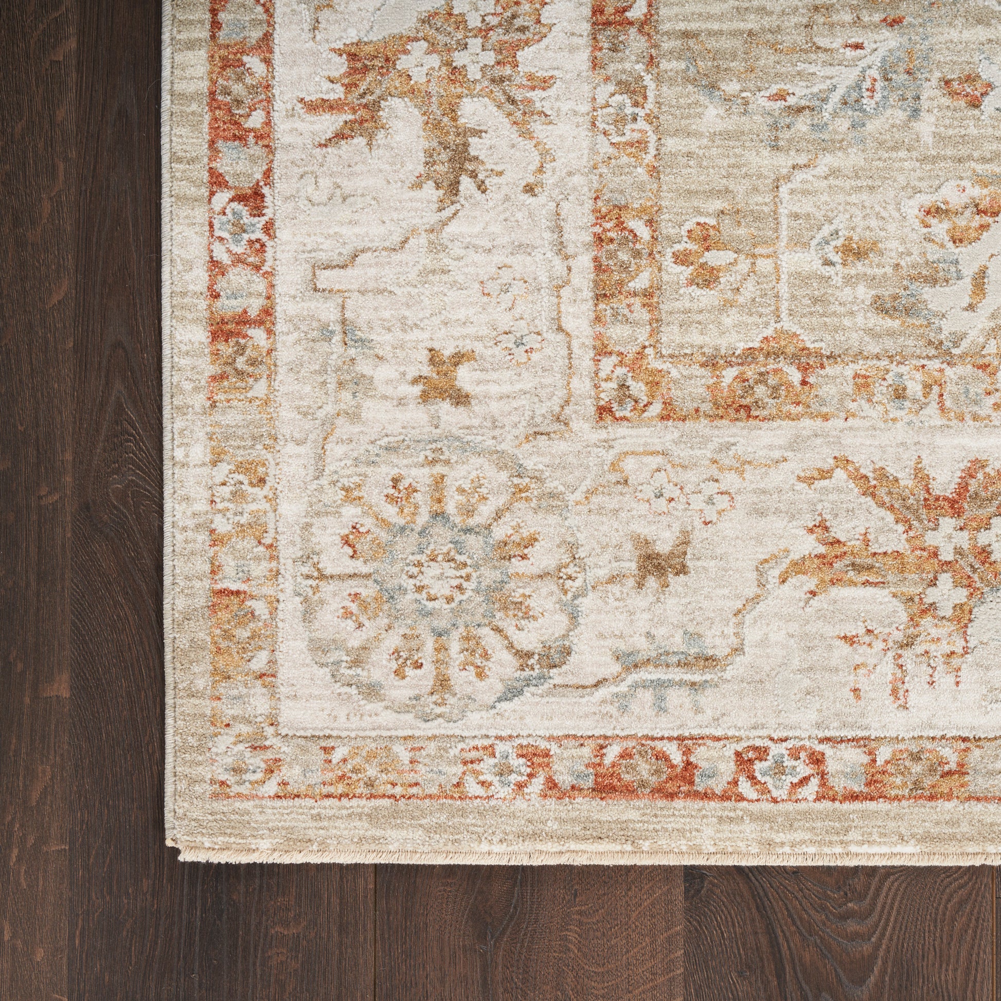 Beige Oriental rug with orange floral border & blue accents on dark wood floor; fringe edge visible.