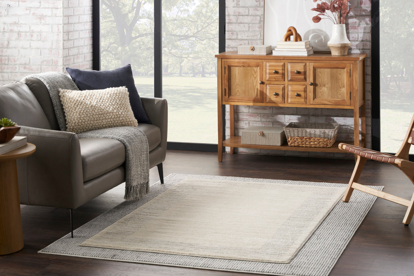 Cozy living room with gray sofa, knit throw, textured pillows, layered beige rug, and a wooden sideboard by large windows.